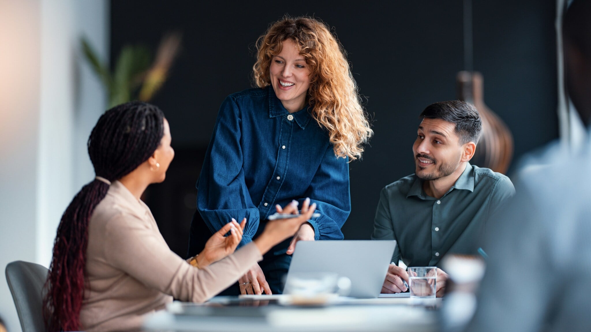 Eine Frau beugt sich über einen Tisch, auf dem ein Laptop steht und an dem zwei weitere Personen sitzen. Eine Frau beugt sich über einen Tisch, auf dem ein Laptop steht und an dem zwei weitere Personen sitzen.