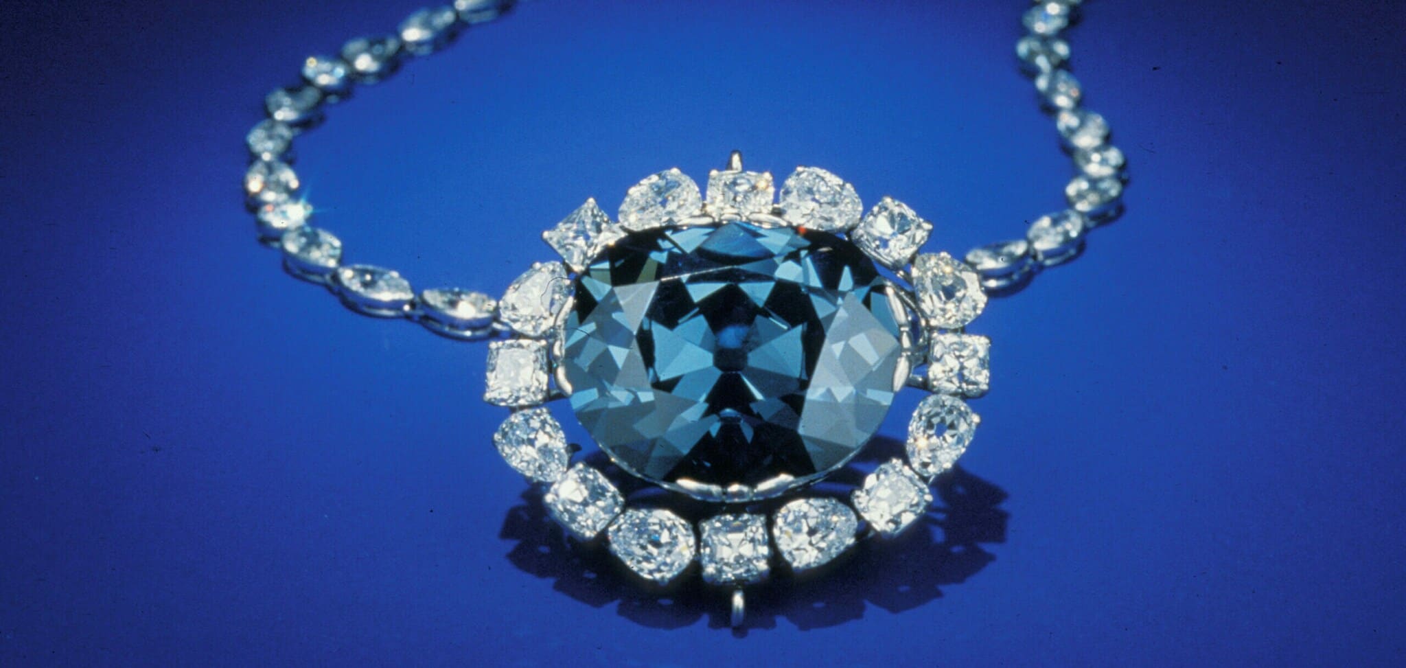 Ein großer, blauer Diamant, eingefasst in kleinere Diamanten an einer Kette auf blauem Untergrund Ein großer, blauer Diamant, eingefasst in kleinere Diamanten an einer Kette auf blauem Untergrund