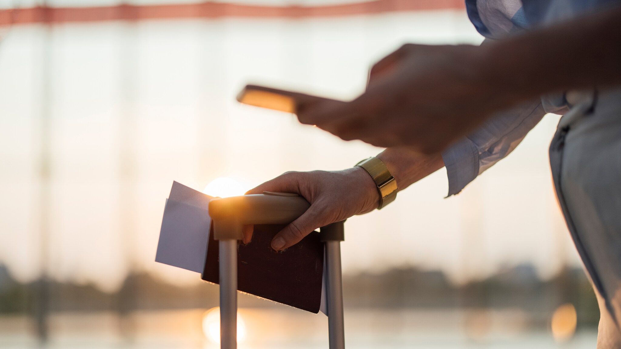 Hand hält einen Reisepass mit Papieren und ein Smartphone über einem Trolleygriff im Flughafen Sonnenuntergang im Hintergrund.