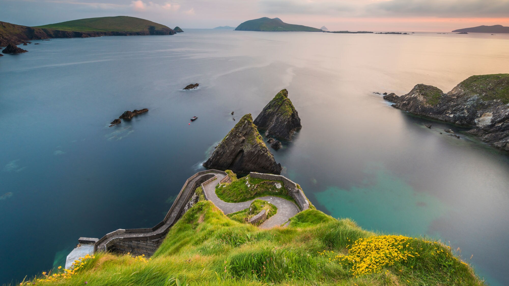 Blick auf eine Küstenlandschaft mit grünem Hügel, steilen Klippen und ruhigem Wasser. Im Hintergrund sind mehrere Inseln und ein Boot sichtbar.