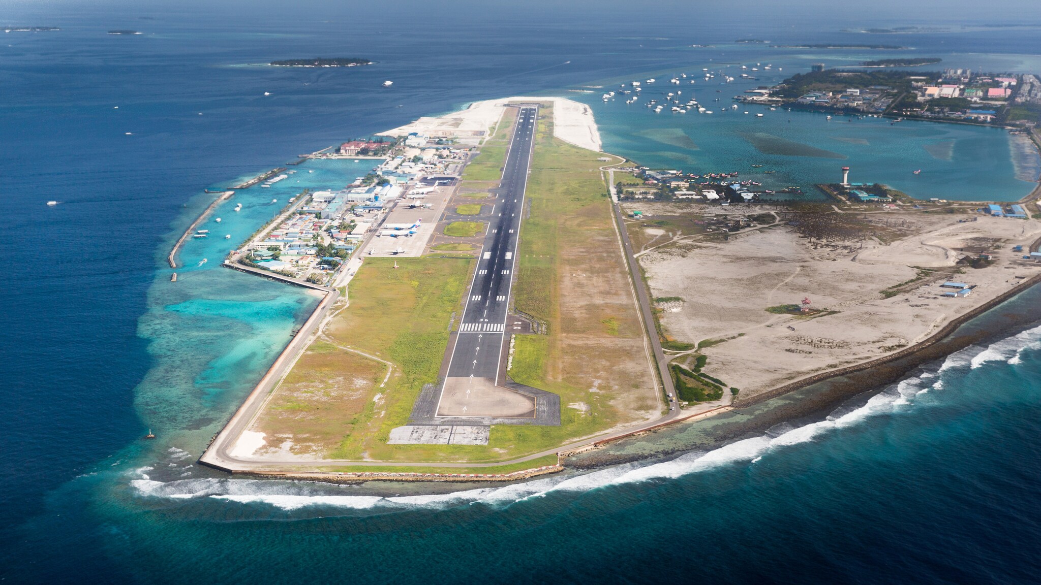 Luftaufnahme eines schmalen Insel-Flughafens mit einer Landebahn, umgeben von Meer und Booten