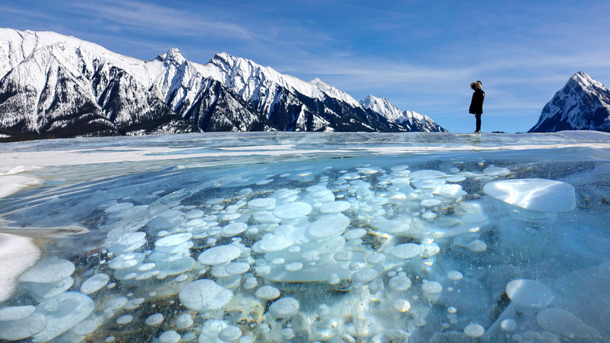 Person steht auf zugefrorenem See mit sichtbaren eingeschlossenen Luftblasen, im Hintergrund schneebedeckte Berge unter blauem Himmel.