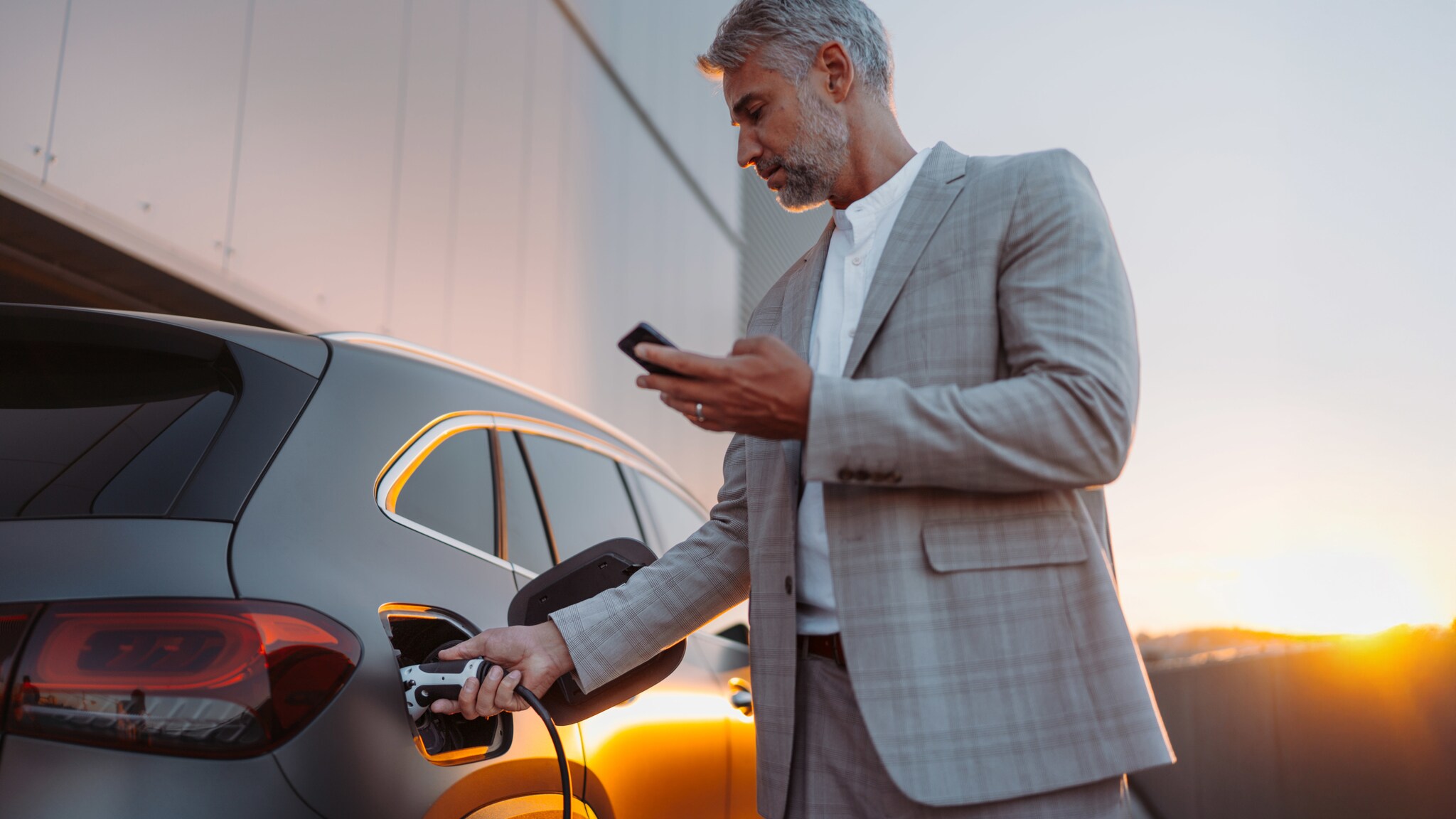 Ein Mann im grauen Anzug und mit einem Smartphone in der Hand steckt den Stecker einer E-Ladesäule in ein Auto.