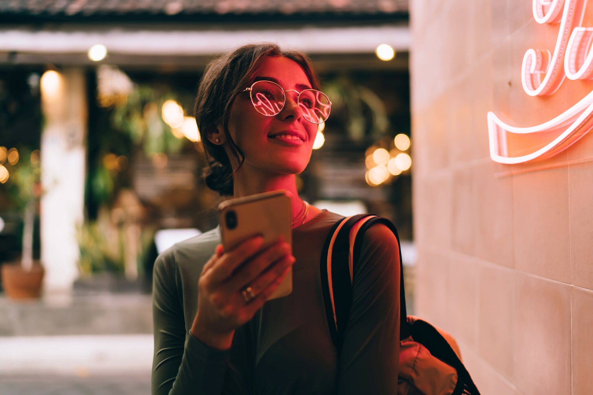 Eine junge Frau hält lächelnd ein Smartphone vor sich in der Hand, in ihrer Brille spiegelt sich der Schriftzug eines roten Logos an einer Wand. Eine junge Frau hält lächelnd ein Smartphone vor sich in der Hand, in ihrer Brille spiegelt sich der Schriftzug eines roten Logos an einer Wand.