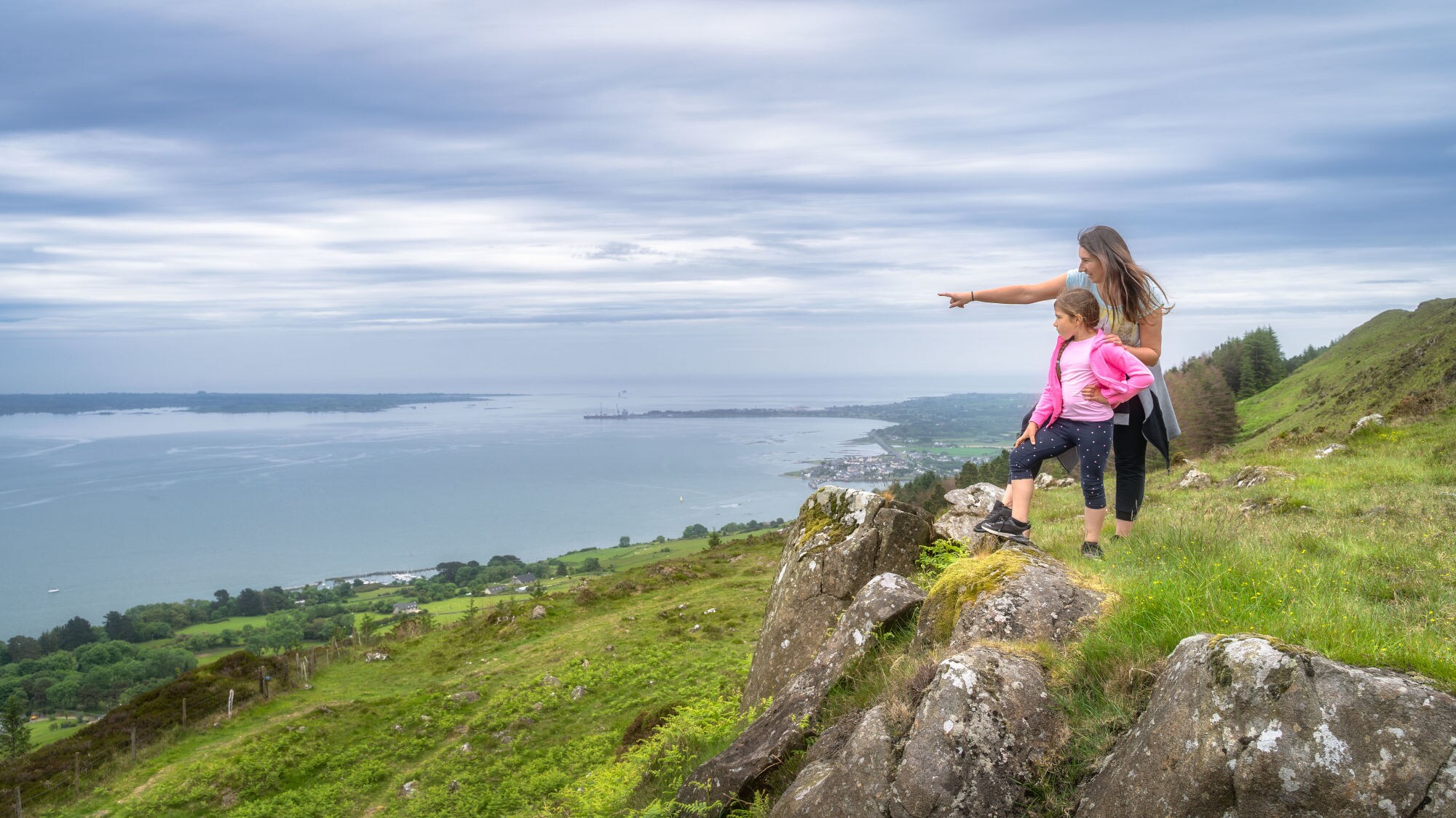 Eine Frau steht mit einem Mädchen auf einem Hügel in einer grünen Graslandschaft an einer Küste und zeigt hinaus aufs Meer. Eine Frau steht mit einem Mädchen auf einem Hügel in einer grünen Graslandschaft an einer Küste und zeigt hinaus aufs Meer.