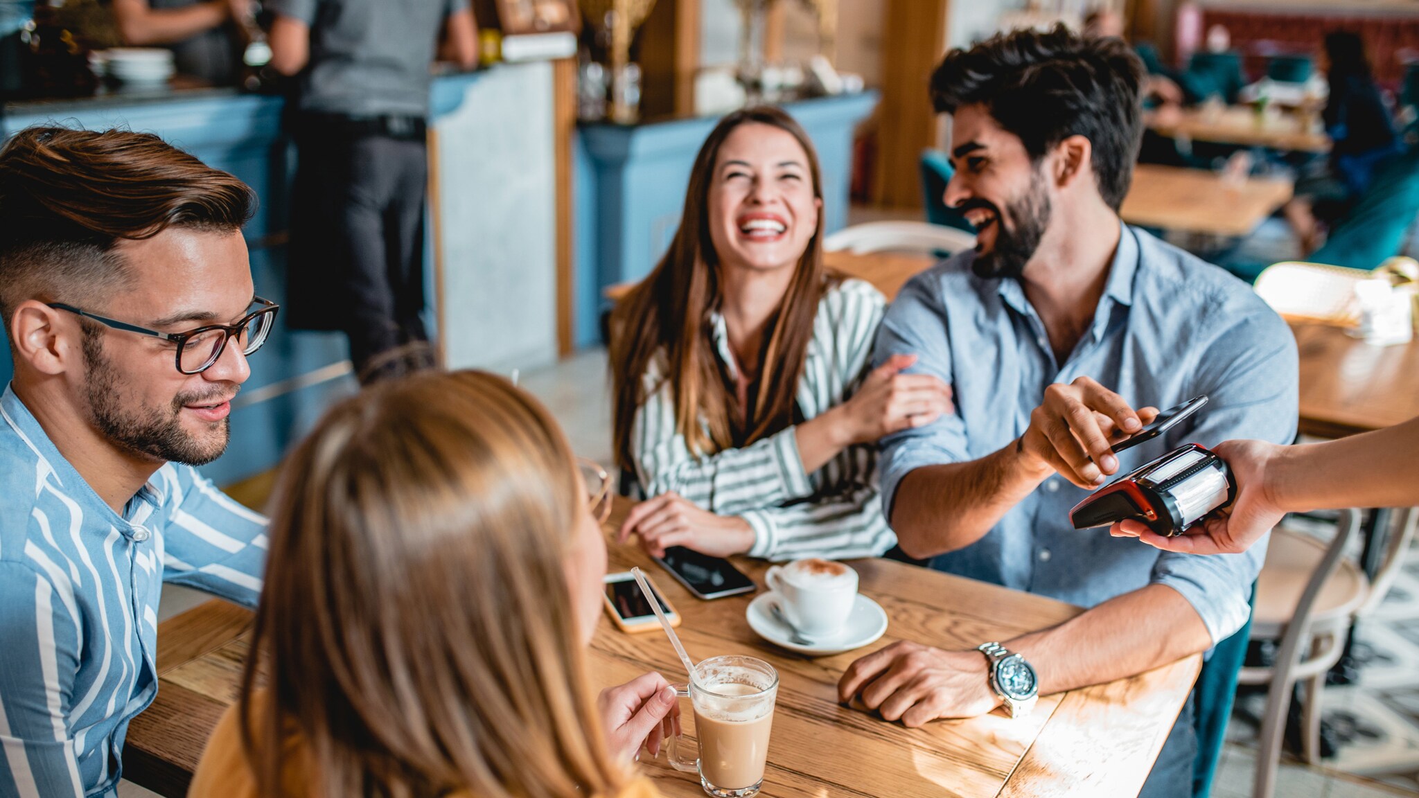 Vier gut gelaunte Personen an einem Tisch in einem Café, eine Person bezahlt kontaktlos mit seinem Smartphone an einem Kartenlesegerät.