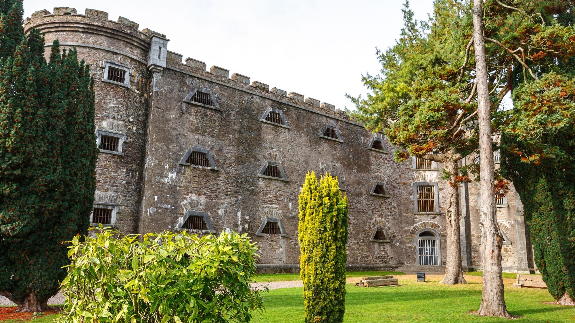 Historisches Gebäude mit steiniger Fassade und Fenster mit Gittern, umgeben von Bäumen und Rasenflächen. Historisches Gebäude mit steiniger Fassade und Fenster mit Gittern, umgeben von Bäumen und Rasenflächen.