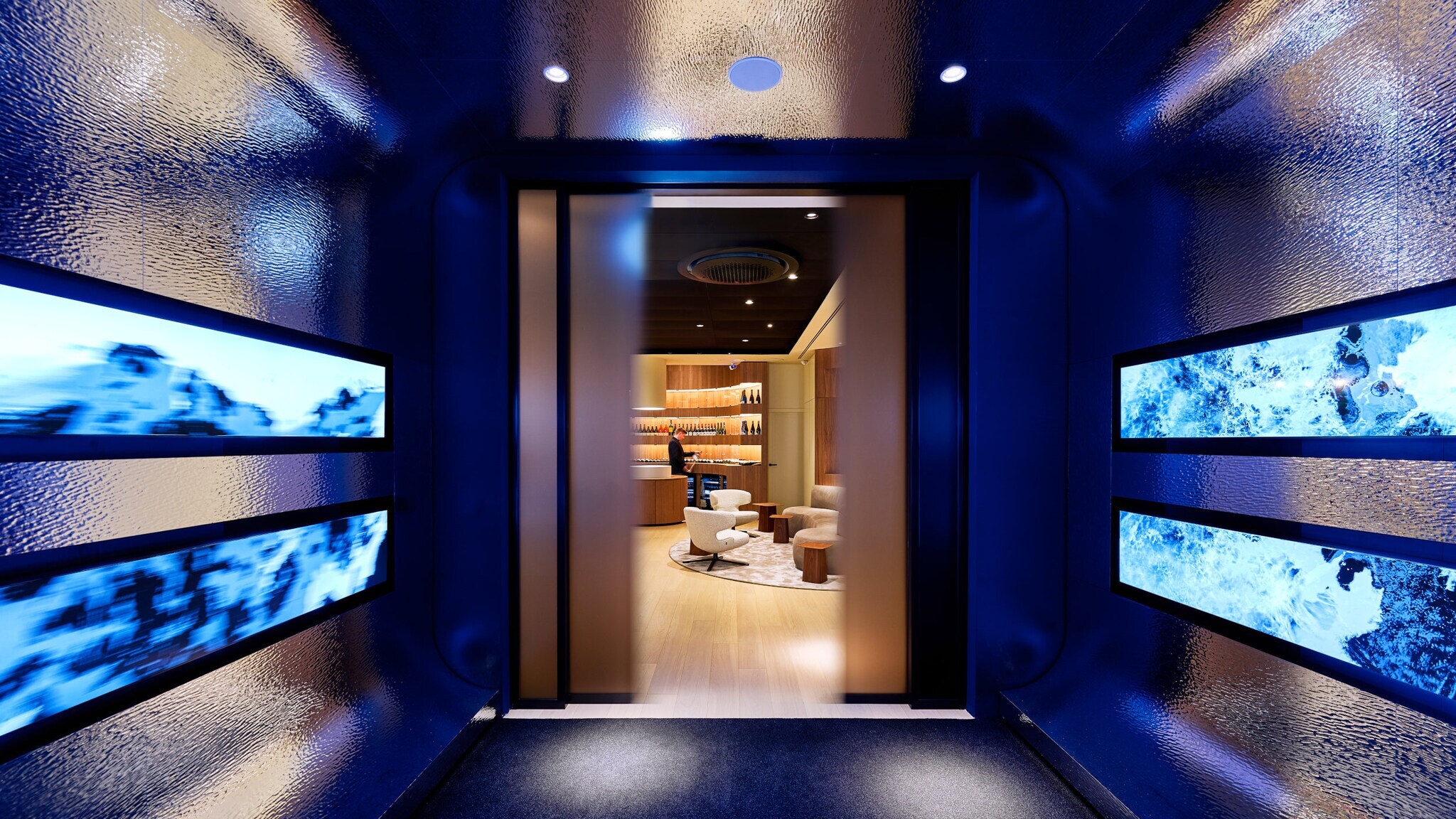 Blick durch einen dunklen, blau beleuchteten Gang mit vier rechteckigen Bildschirmen an den Seiten auf einen hellen, modernen Raum mit Sesseln und Regalen.