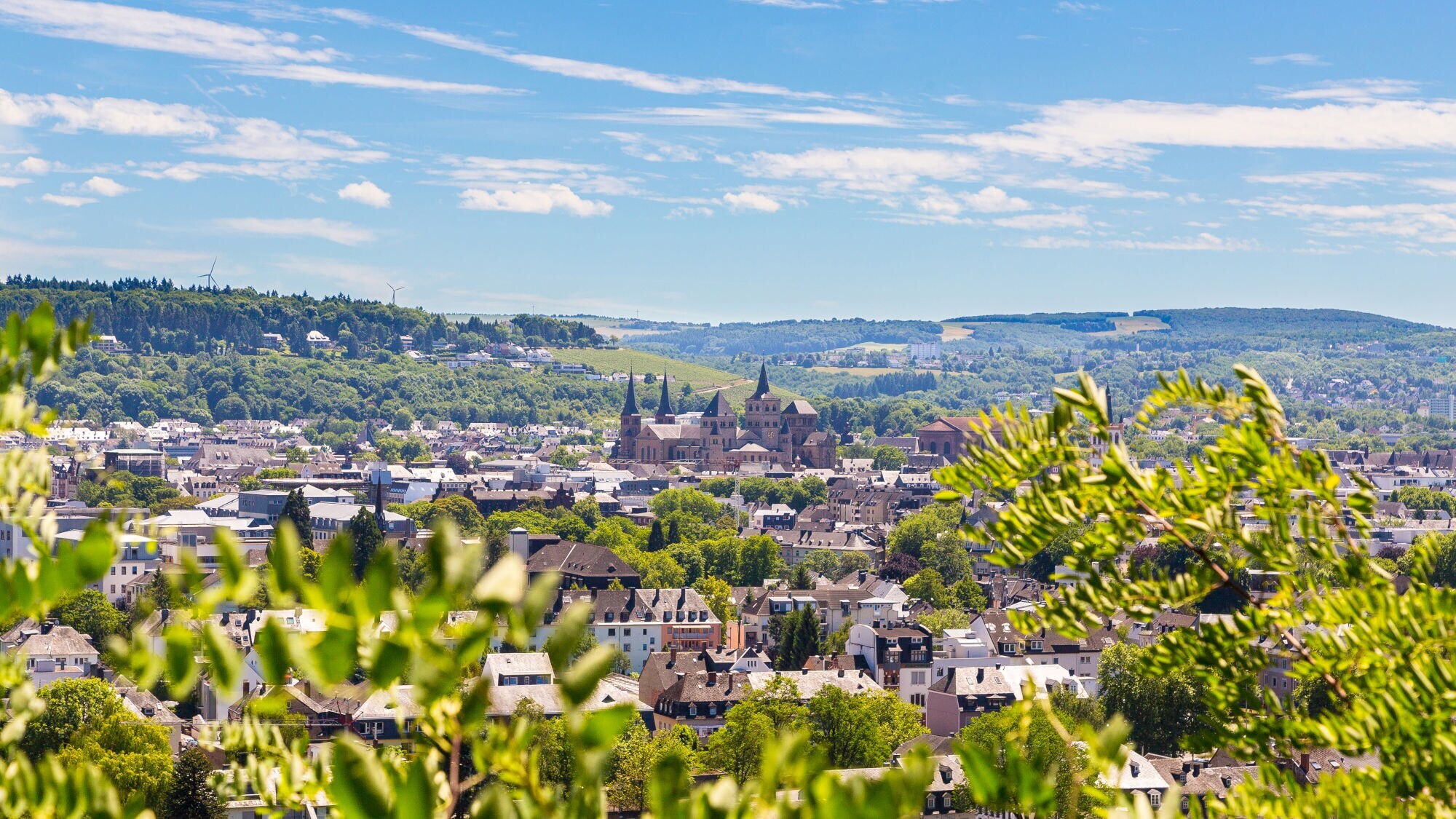 Blick auf Trier mit der Porta Nigra und umliegenden Gebäuden, im Vordergrund grüne Blätter, im Hintergrund bewaldete Hügel unter blauem Himmel mit Wolken.