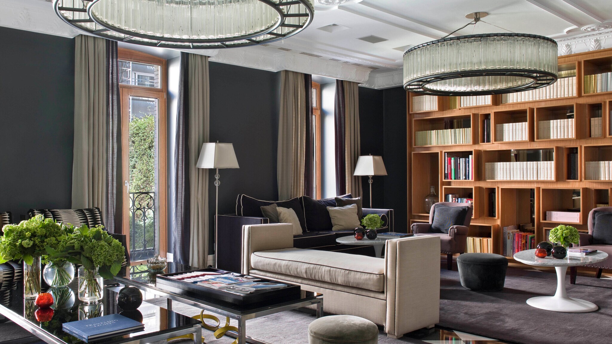Hotelbibliothek im Hotel Unico Madrid mit zwei großen, runden Kronleuchtern, einem beigen Sofa, einem Glastisch und einer Bücherwand. Pflanzen und Dekorationen auf dem Tisch.