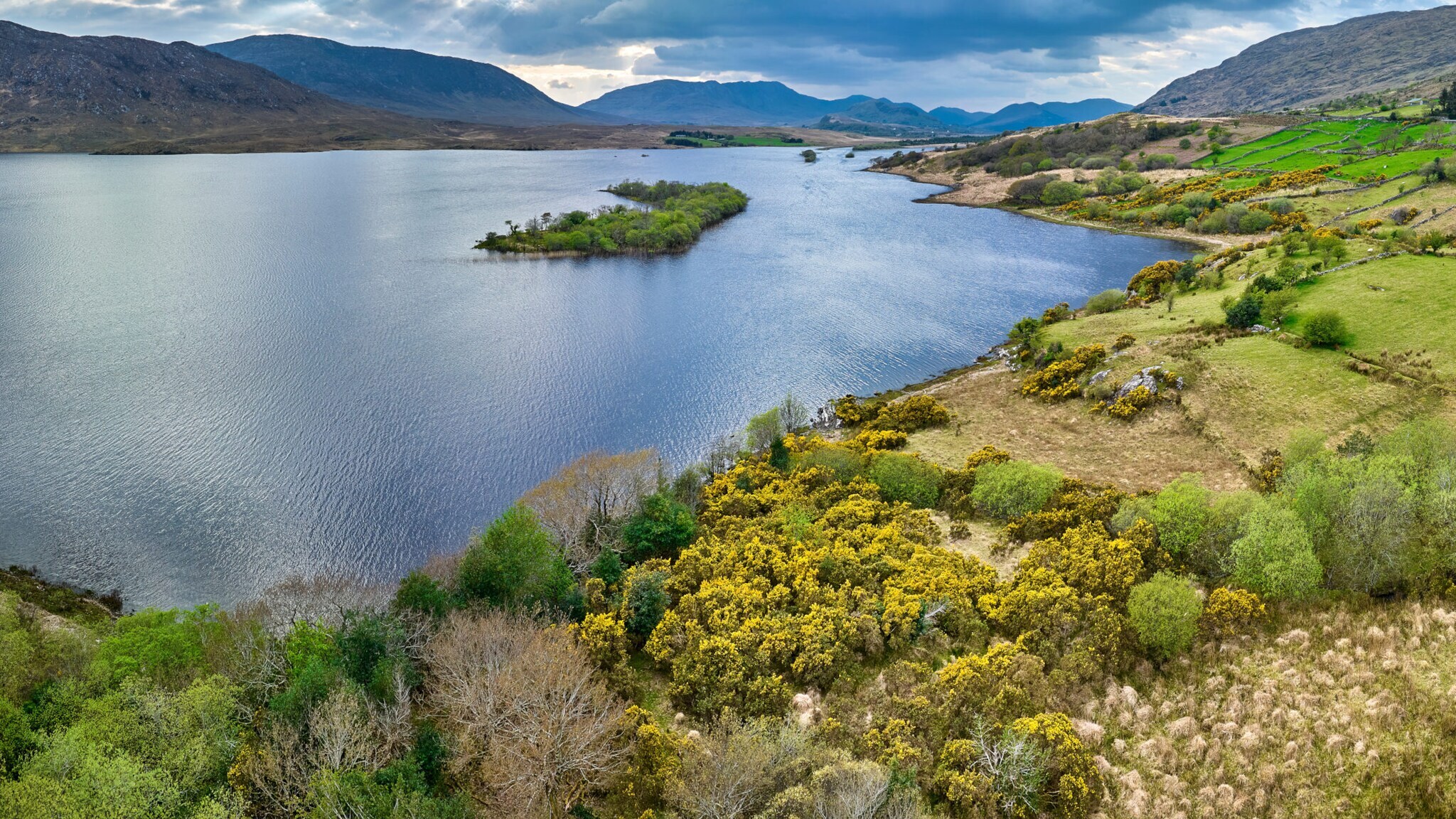 Panorama einer Landschaft mit einem ruhigen See, umgeben von Hügeln, Bäumen und grünen Feldern. Im Hintergrund sind Berge sichtbar. Panorama einer Landschaft mit einem ruhigen See, umgeben von Hügeln, Bäumen und grünen Feldern. Im Hintergrund sind Berge sichtbar.