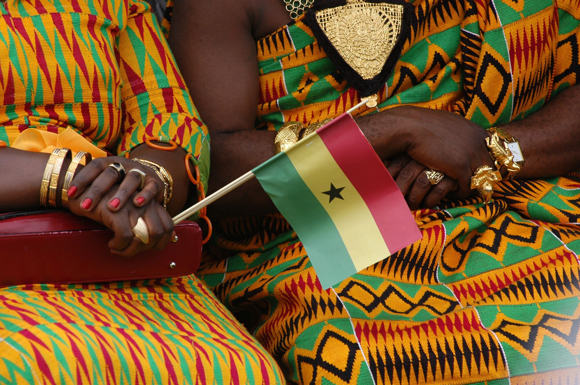 Oberkörper zweier sitzender Frauen in traditionellen ghanaischen Kleidern aus buntem Kente-Stoff und Goldschmuck, davor eine kleine ghanaische Flagge aus Papier in einer Hand gehalten.