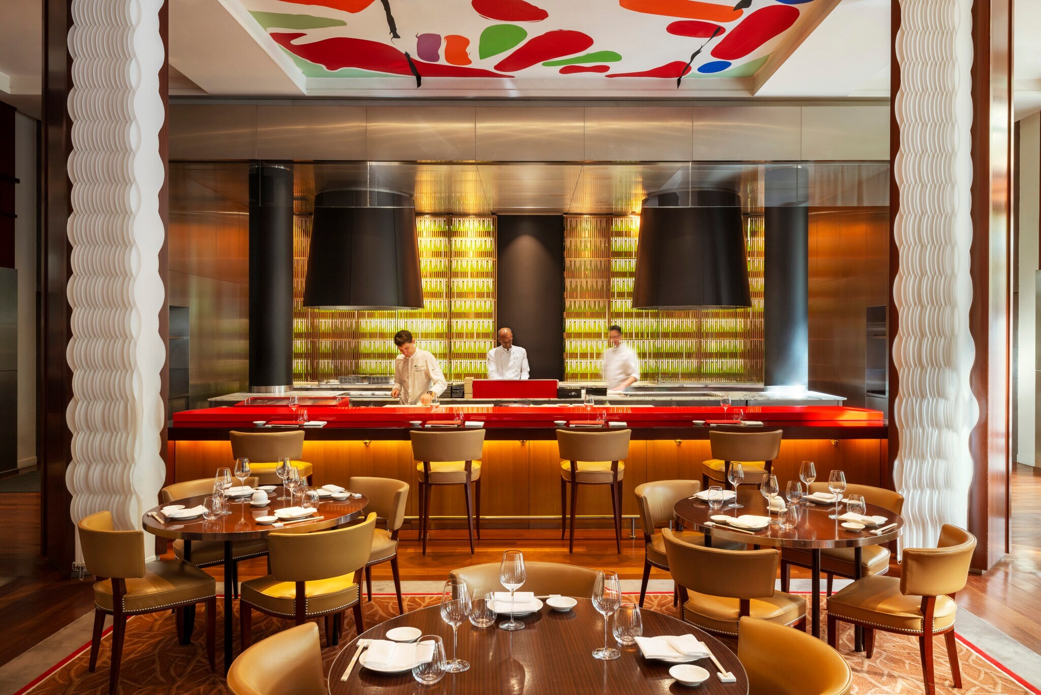 Interieur eines elegant-modernen Restaurants mir runden gedeckten Holztischen und offenem Tresen, in dem drei Köche stehen.