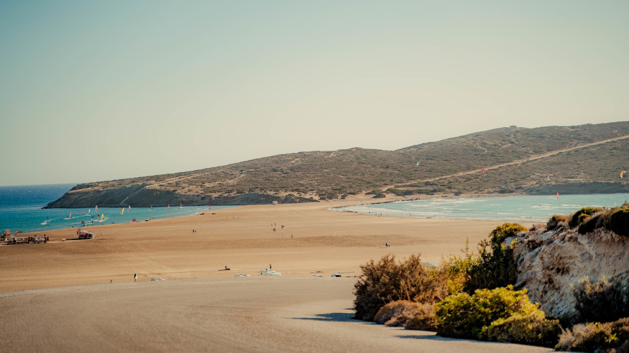 Ein sonniger Tag am Strand von Prasonisi. Die schmale Sandbank führt zur kleinen hügeligen Halbinsel und auf dem Wasser sowie in der Luft tummeln sich bunte Surfer:innen und Kitesurfer:innen.