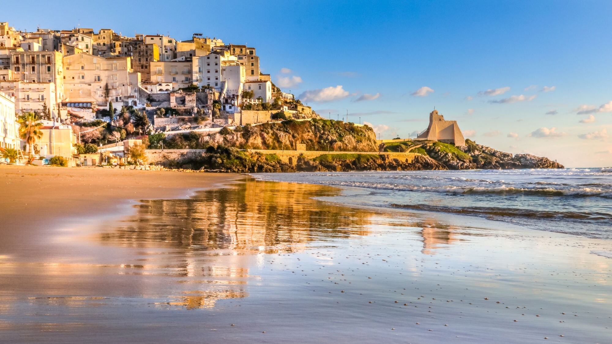 Strand mit nassem Sand, der die Häuser von Sperlonga und einen Turm auf einer Klippe im Sonnenlicht spiegelt.