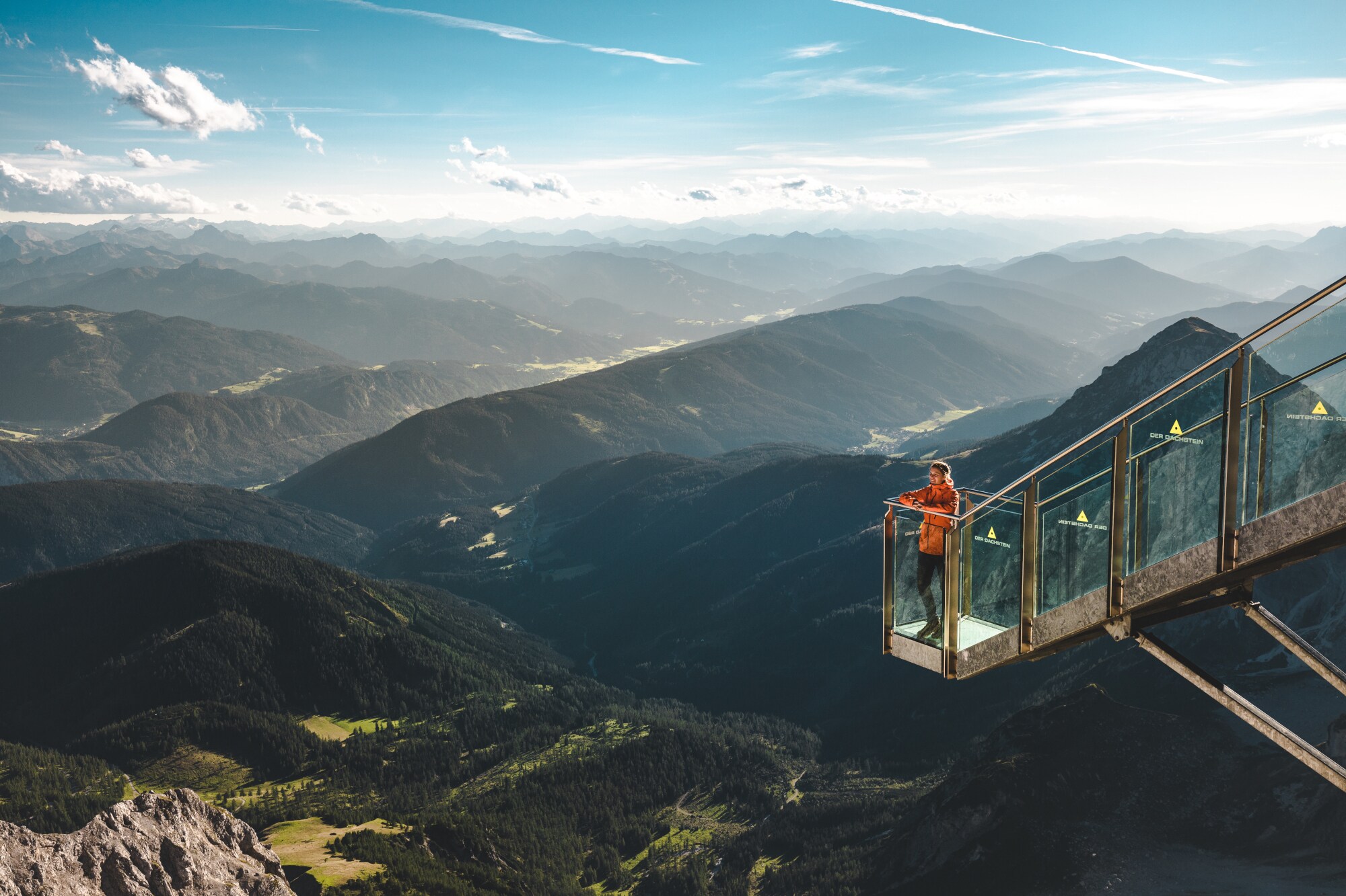 Eine Person steht auf einer gläsernen Aussichtsplattform vor Berglandschaft. Eine Person steht auf einer gläsernen Aussichtsplattform vor Berglandschaft.