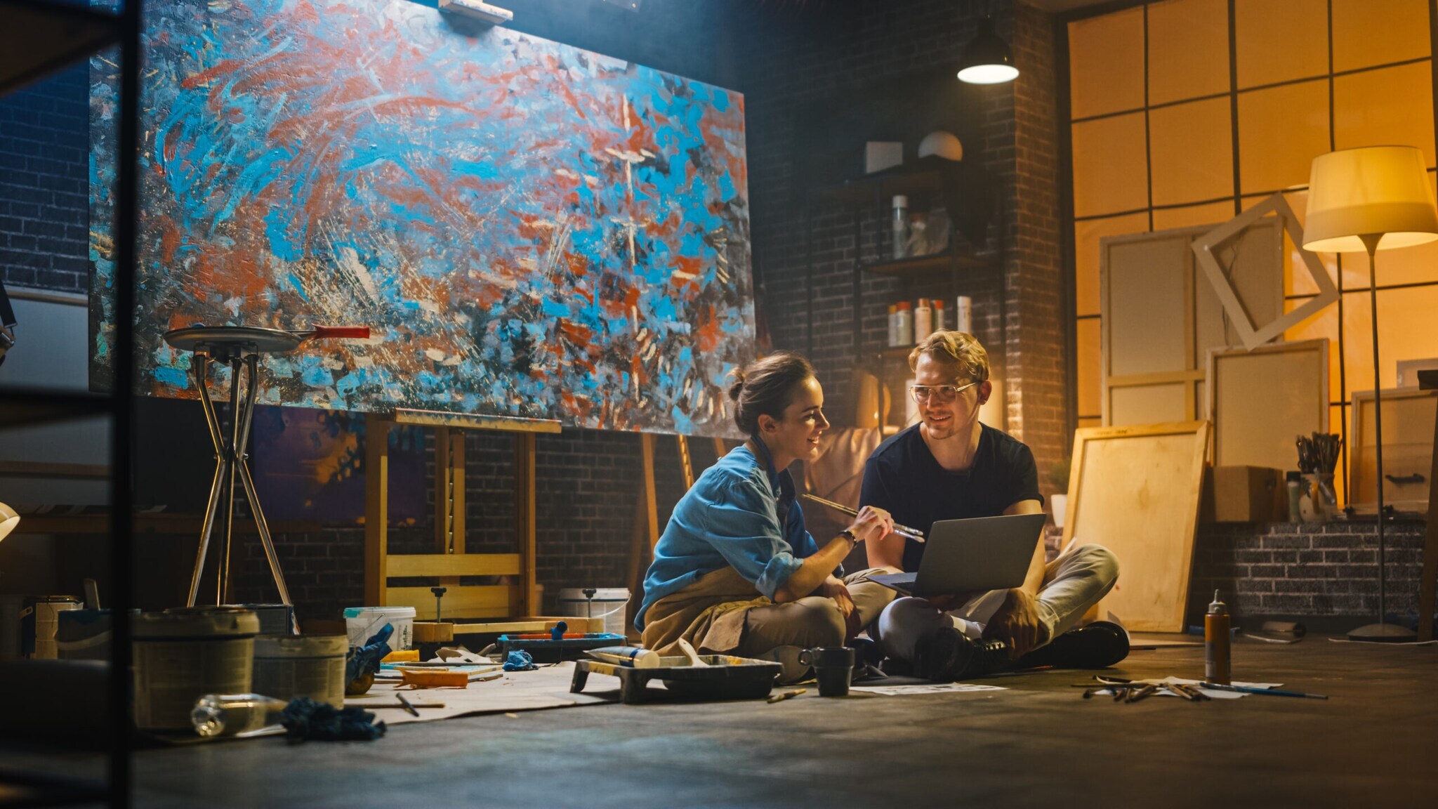 Ein junges Pärchen sitzt in einem Atelier vor einem Laptop und unterhält sich