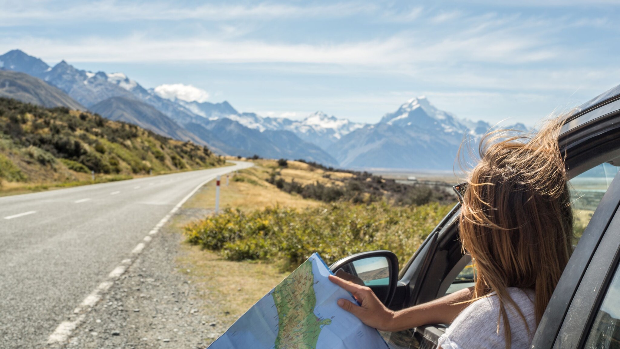 Frau schaut aus Autofenster auf eine Landkarte mit Blick auf eine offene Straße und schneebedeckte Berge im Hintergrund. Frau schaut aus Autofenster auf eine Landkarte mit Blick auf eine offene Straße und schneebedeckte Berge im Hintergrund.