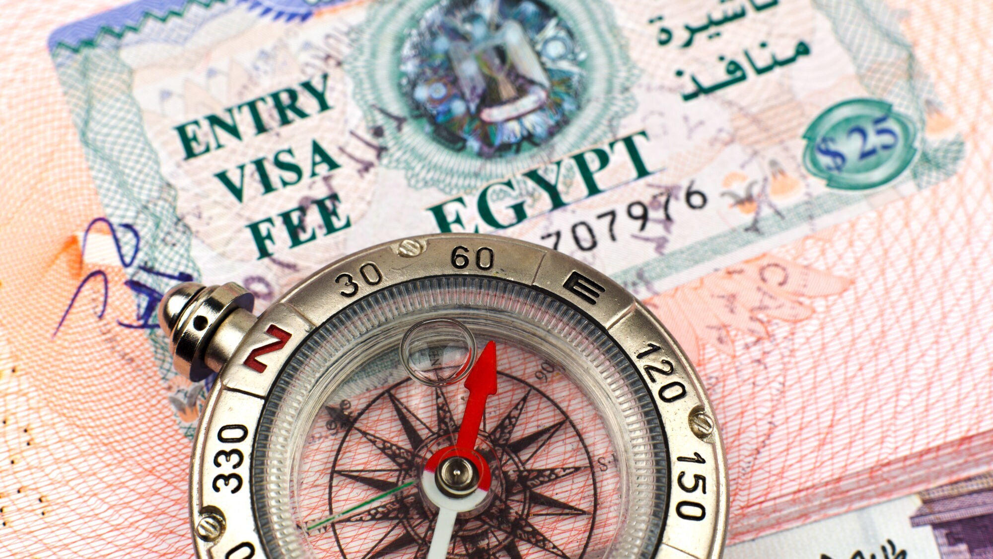 Nahaufnahme eines Kompasses mit rotem Zeiger vor einem ägyptischen Visum mit dem Text 'ENTRY VISA FEE EGYPT' und arabischer Schrift.