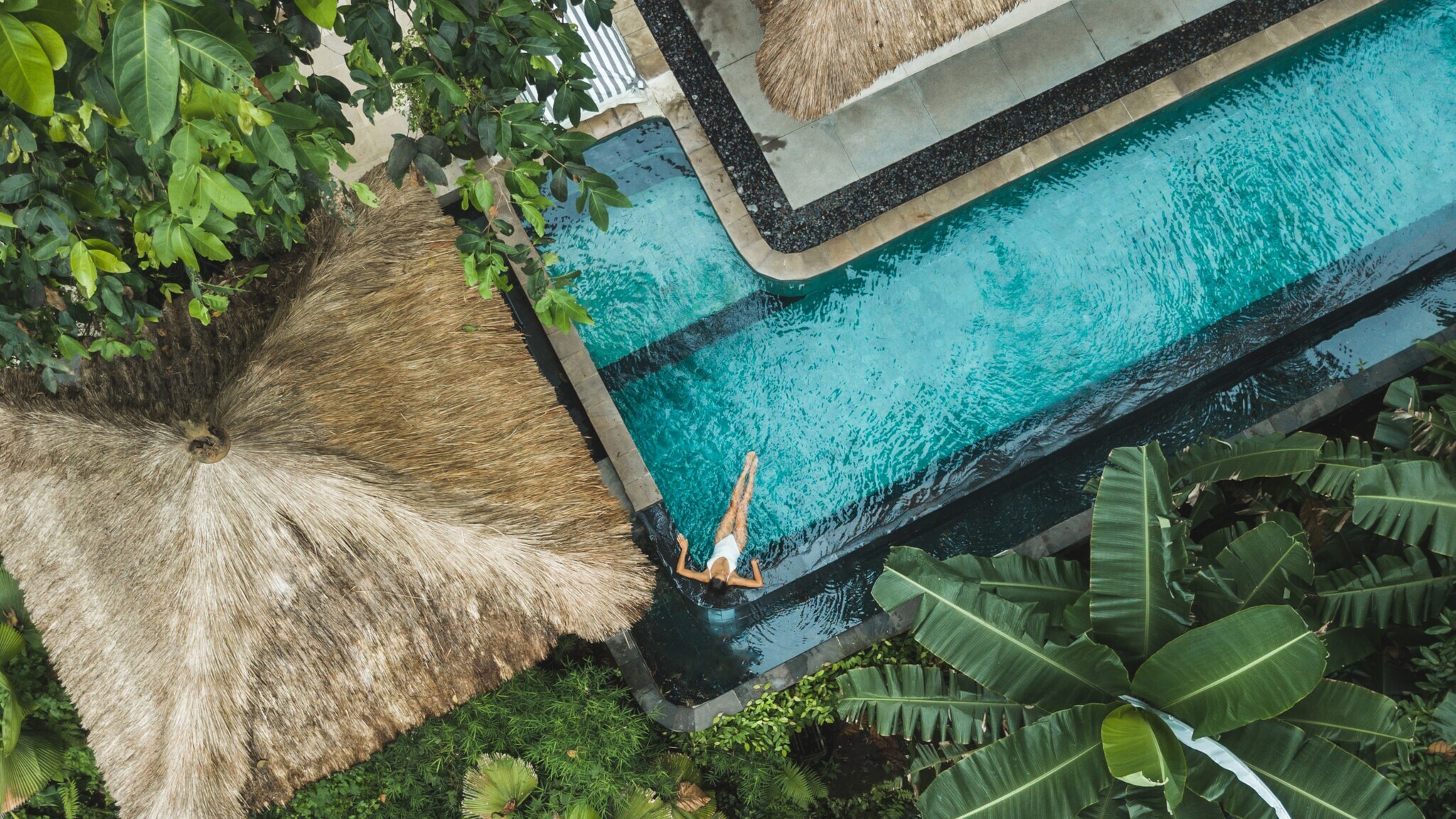 Eine Person schwimmt in einem blauen Pool, umgeben von tropischen Pflanzen und einem Strohdach. Der Blick ist von oben.