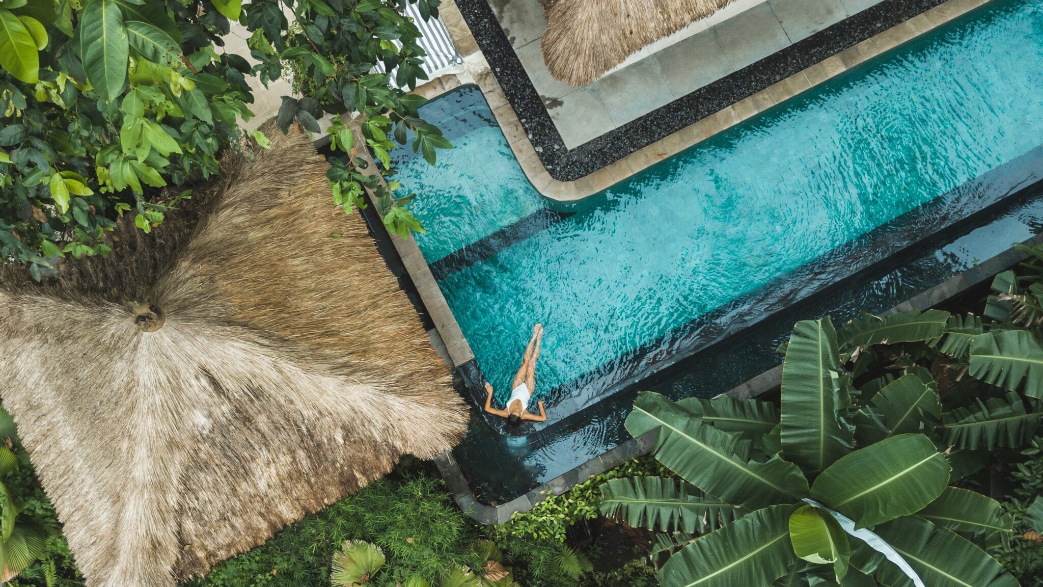 Eine Person schwimmt in einem blauen Pool, umgeben von tropischen Pflanzen und einem Strohdach. Der Blick ist von oben. Eine Person schwimmt in einem blauen Pool, umgeben von tropischen Pflanzen und einem Strohdach. Der Blick ist von oben.