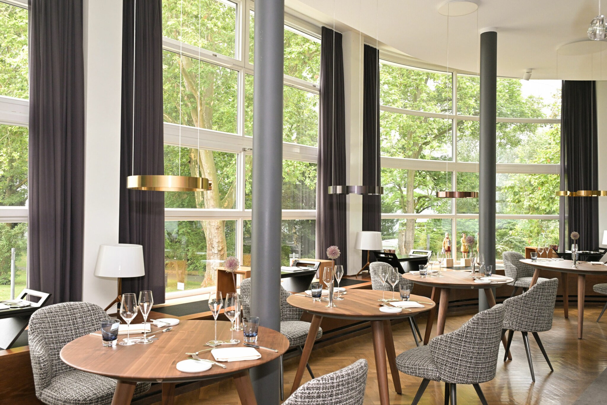 Ein lichtdurchfluteter Saal mit eingedeckten Holztischen und hohe, bodentiefe Fenster