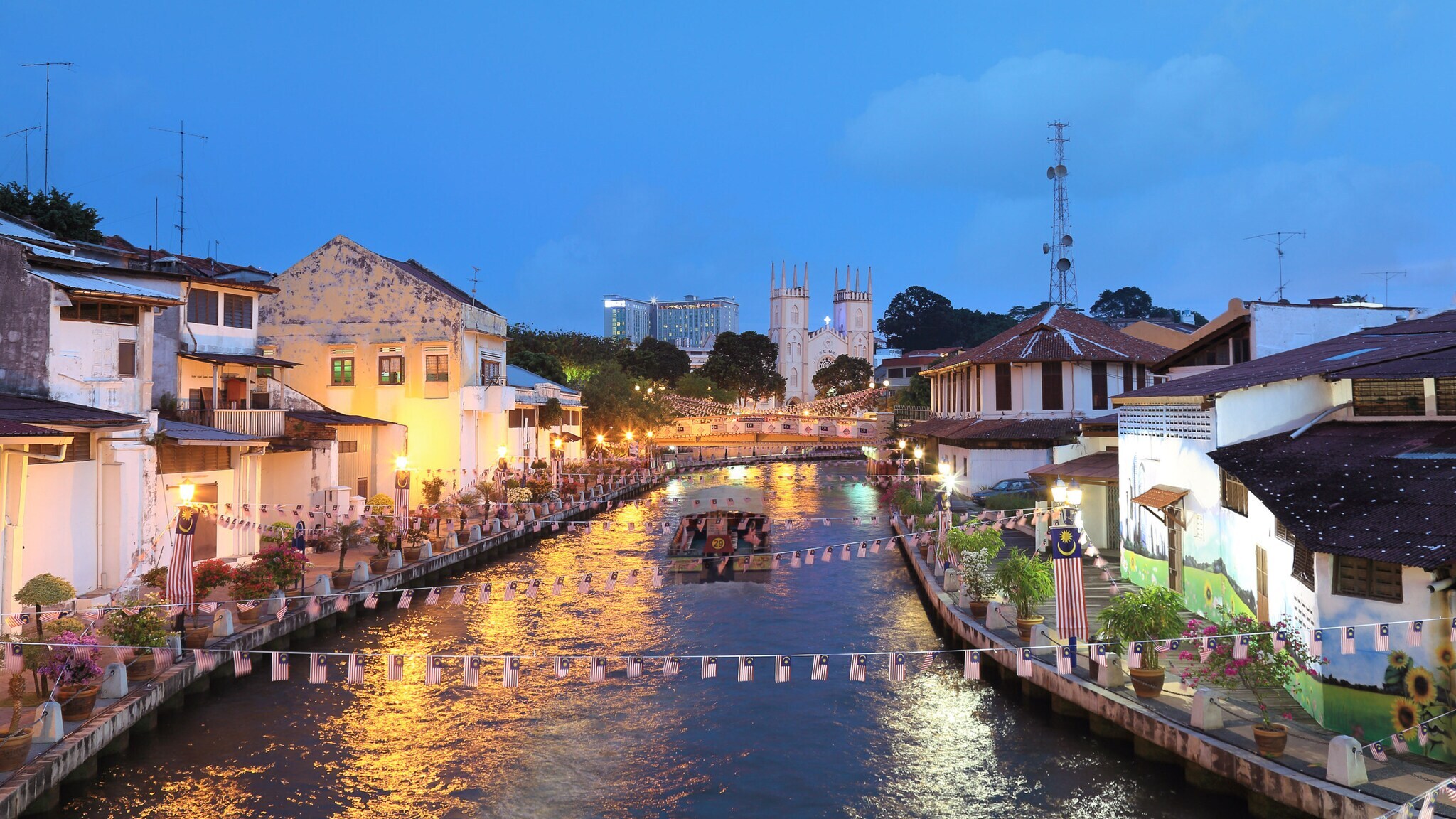 Ein Ausflugsboot fährt über einen mit Girlanden geschmückten Kanal durch eine Stadt mit flachen Häusern in Uferlage am Abend, im Hintergrund steht eine Kirche mit Zwillingstürmen.