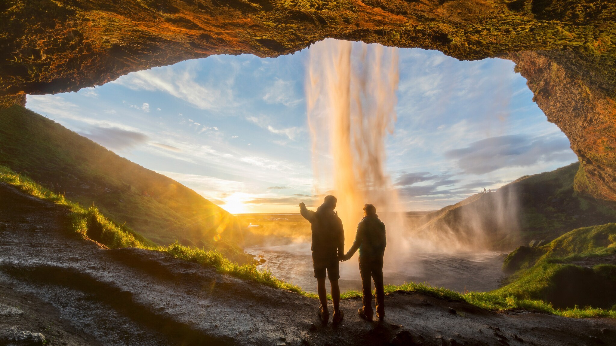 Zwei Personen stehen in einer Höhle und blicken auf einen Wasserfall bei Sonnenuntergang.