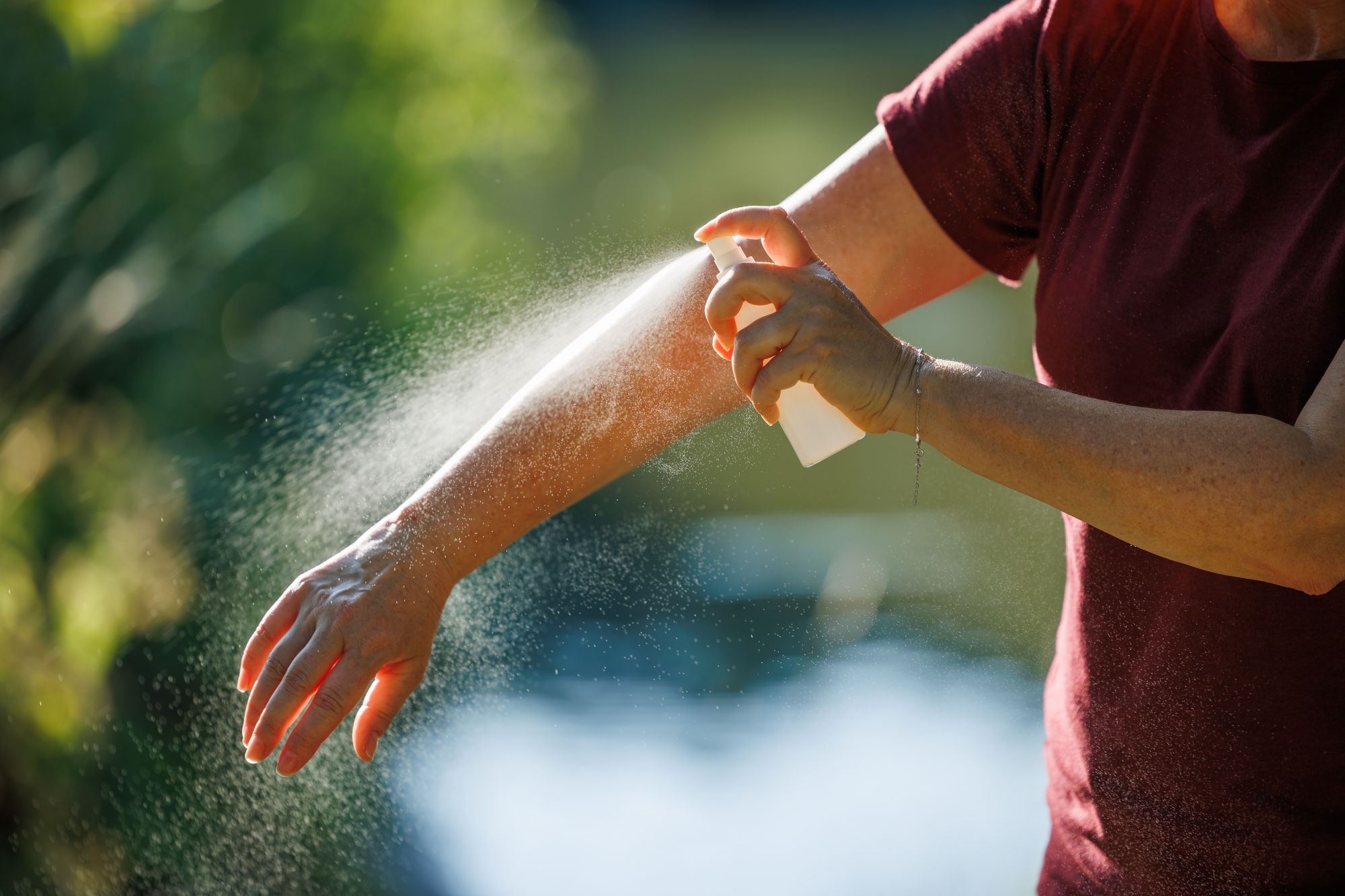 Ein Mann im T-Shirt besprüht seinen Unterarm mit Flüssigkeit aus einer Plastikflasche bei Sonnenschein.