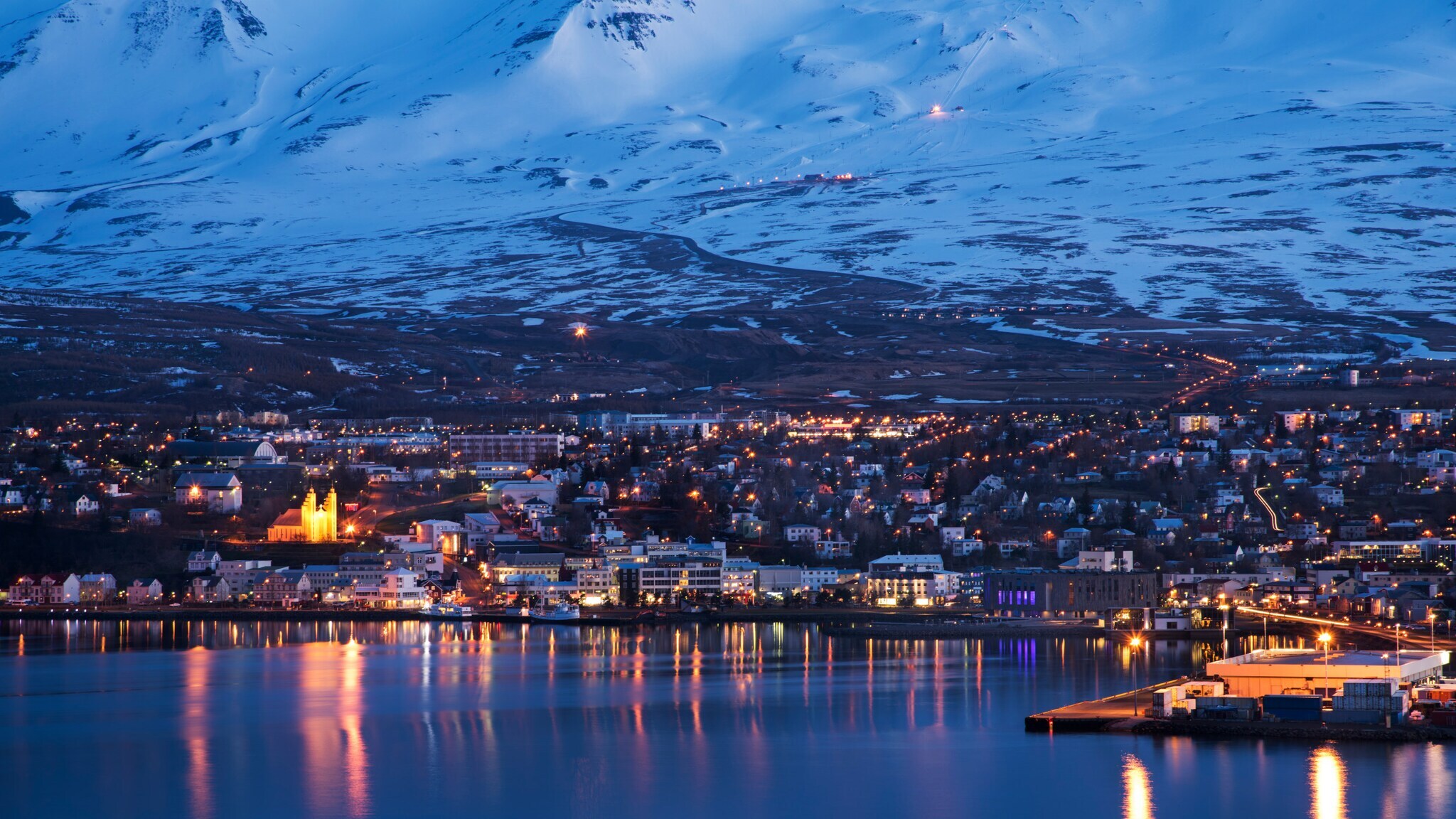 Stadt am Ufer eines Gewässers bei Nacht mit beleuchteten Gebäuden und schneebedeckten Bergen im Hintergrund