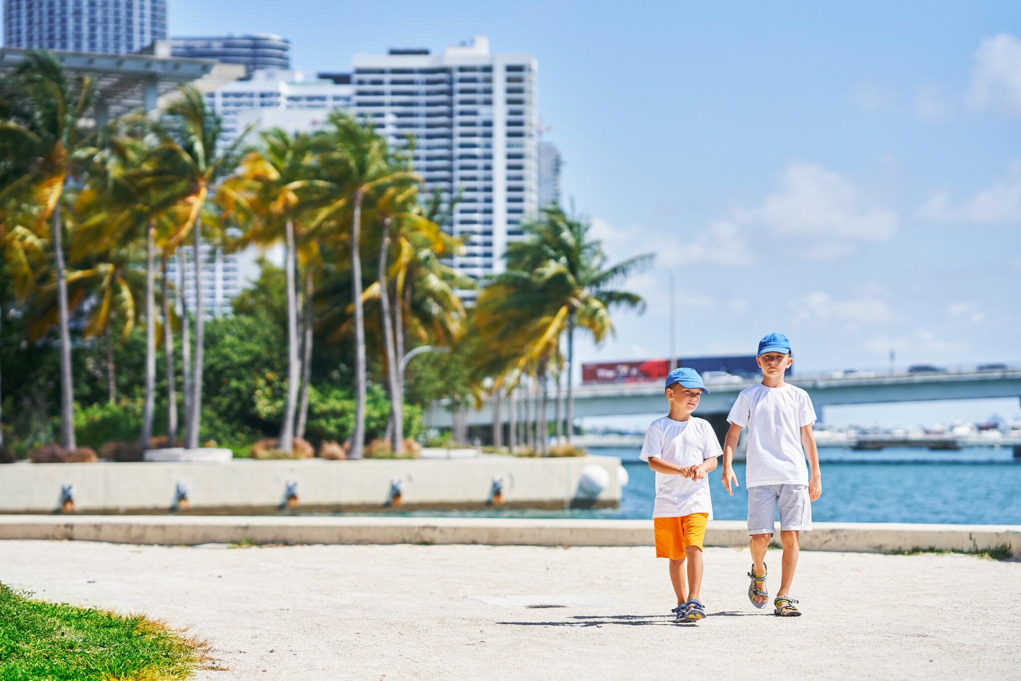 Zwei Jungen mit Schirmmützen spazieren an der Küste Miamis, im Hintergrund Palmen, Hochhäuser und das Meer.