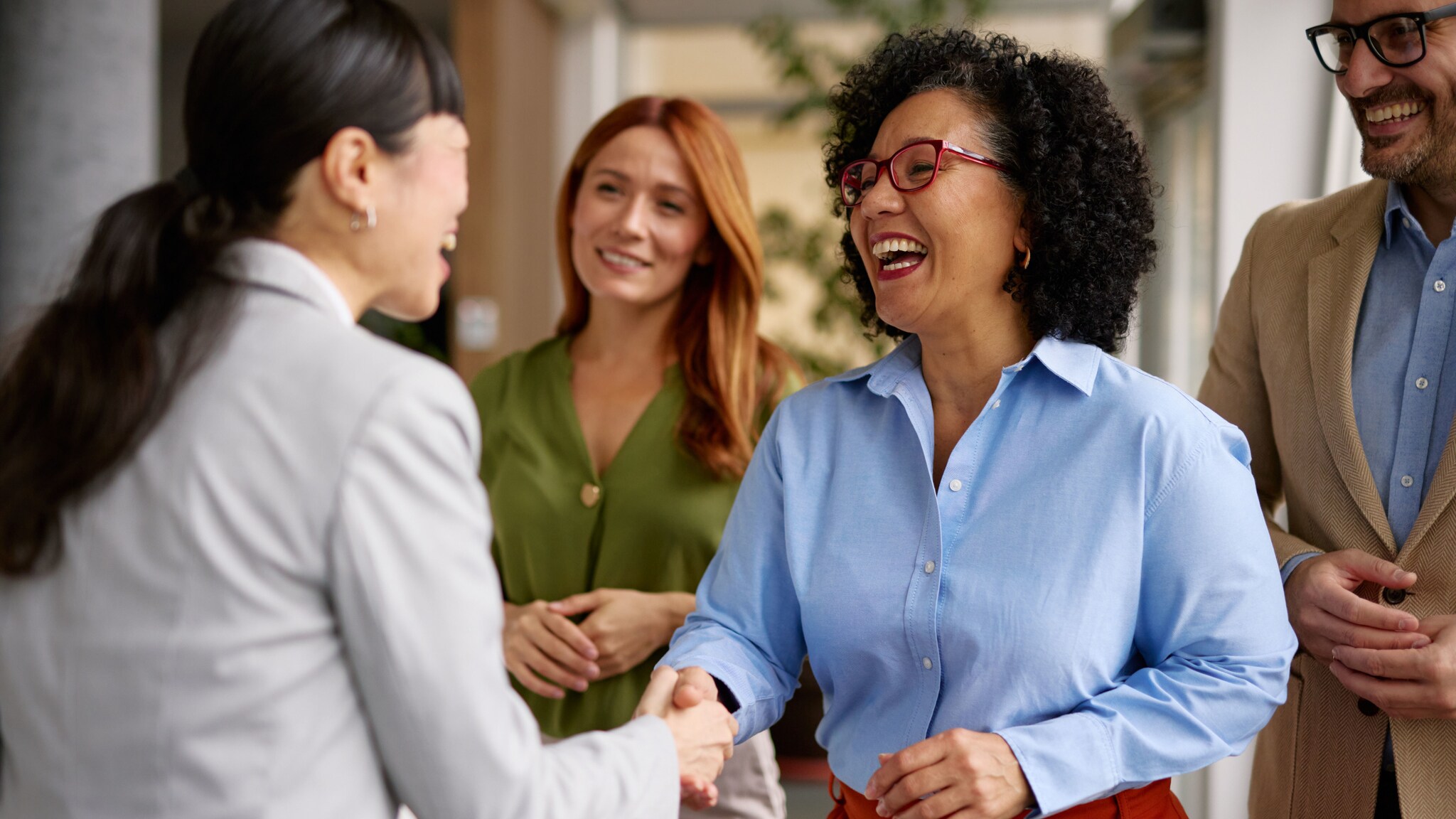 Vier Personen stehen beieinander in einem Büro, im Vordergrund geben sich zwei lachende Frau die Hand. Vier Personen stehen beieinander in einem Büro, im Vordergrund geben sich zwei lachende Frau die Hand.