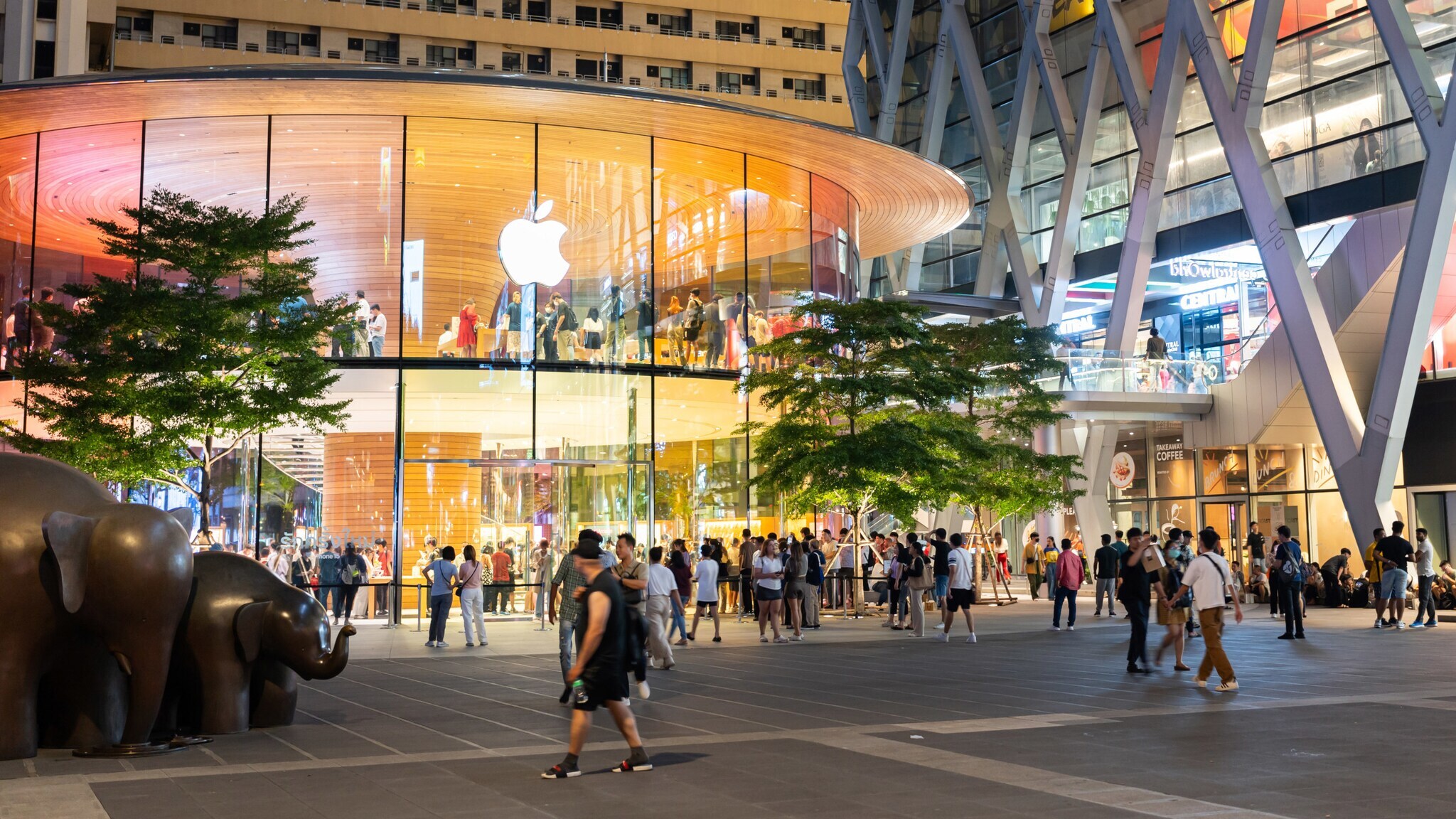 Personen in und auf einem Platz vor einem modernen, erleuchteten Gebäude mit Apple-Logo bei Dunkelheit.