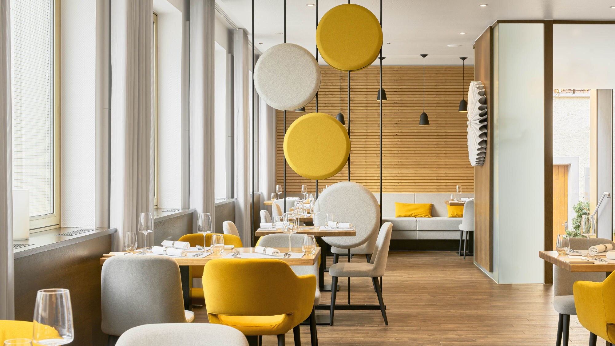 Modern eingerichteter Gastraum mit gelben und grauen Stühlen, Holzwänden und runden, schwebenden Akustikelementen in Gelb und Grau.