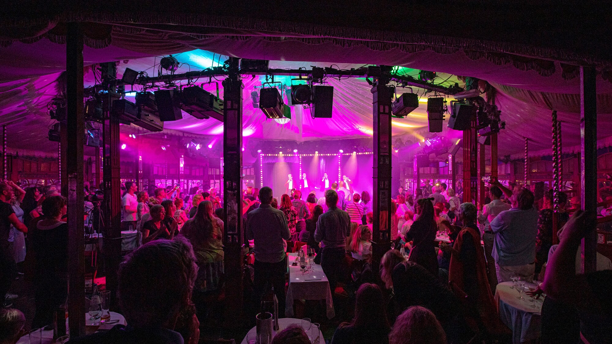 Eine Menschenmenge steht in einem Zelt mit bunten Lichtern. Auf der Bühne sind mehrere Personen sichtbar, die performen. Tische sind im Vordergrund. Eine Menschenmenge steht in einem Zelt mit bunten Lichtern. Auf der Bühne sind mehrere Personen sichtbar, die performen. Tische sind im Vordergrund.