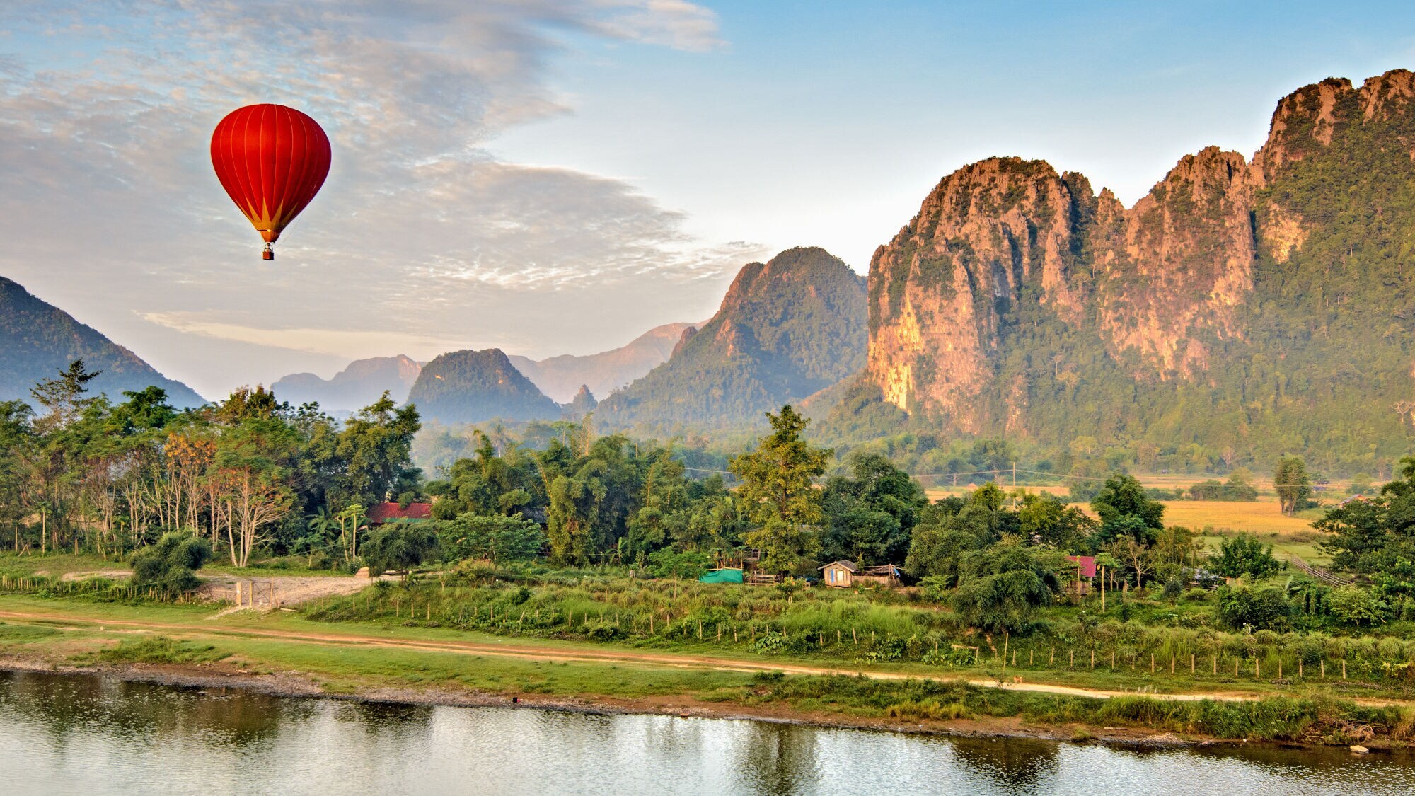 Ein roter Heißluftballon schwebt über einer grünen Landschaft mit Bergen und einem ruhigen Fluss.