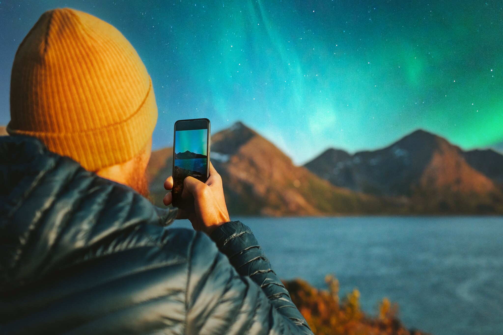 Ein Mann mit gelber Mütze hält ein Smartphone in der Hand und fotografiert damit Polarlichter.