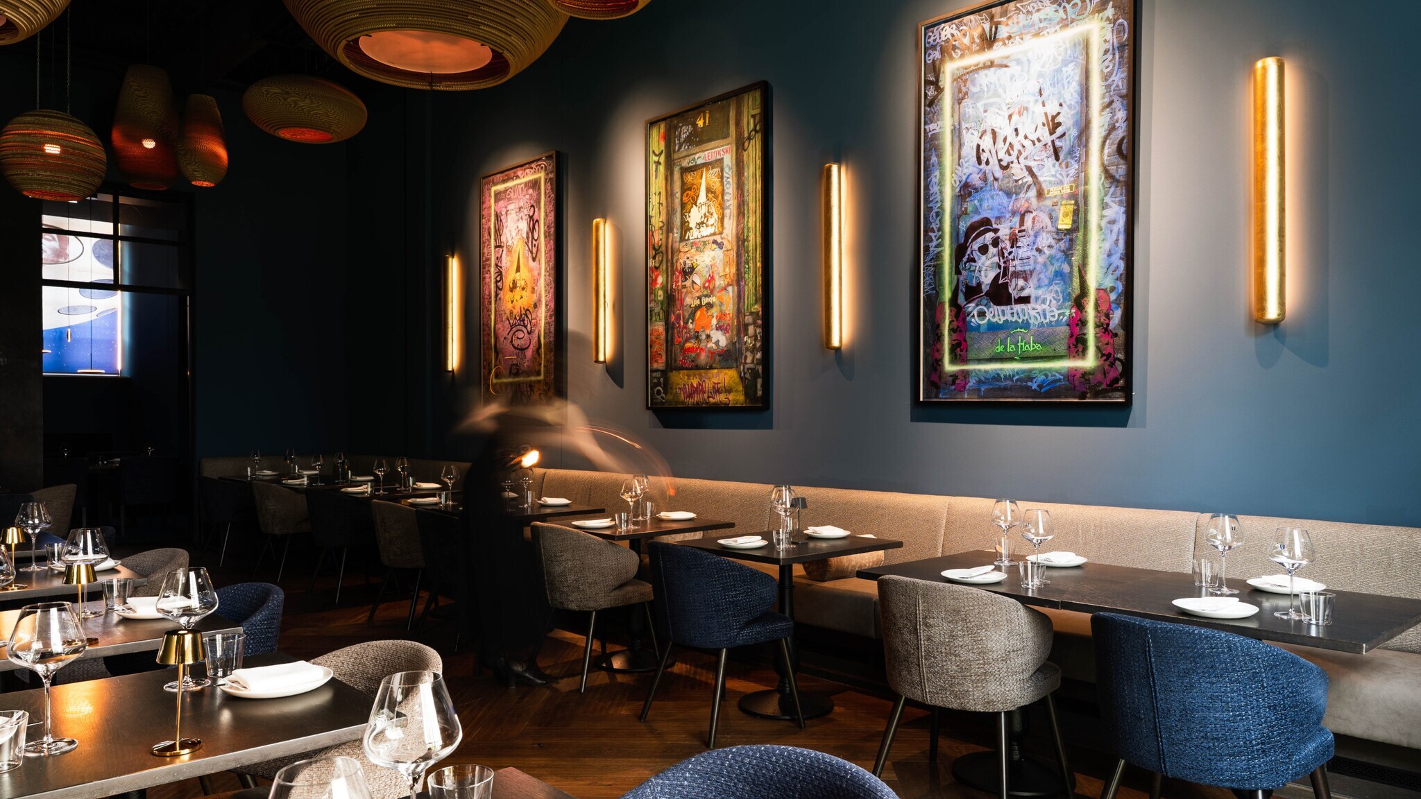Modern eingerichteter Gastraum mit gedeckten Tischen, Stühlen in Grau und Blau, beleuchteten Wandbildern und warmem Licht an den Wänden.
