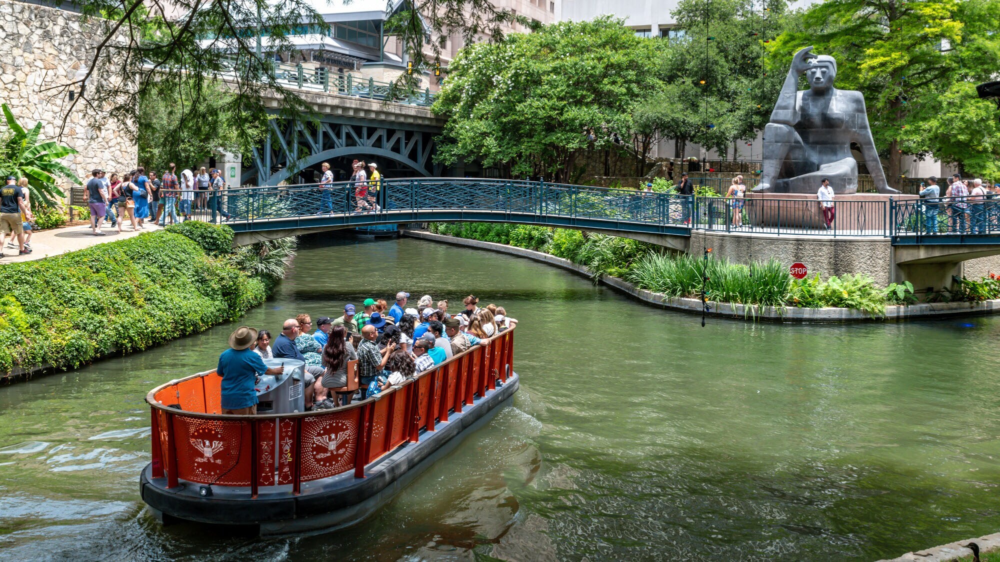 Ausflugsboot auf einer begrünten Wasserstraße vor einer Brücke mit Steinskulptur am im Stadtzentrum San Antonios. Ausflugsboot auf einer begrünten Wasserstraße vor einer Brücke mit Steinskulptur am im Stadtzentrum San Antonios.