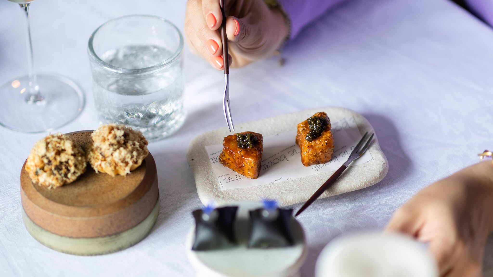 Eine Hand führt eine Gabel zu einem kunstvoll angerichteten Dessert auf einem weiß eingedeckten Restauranttisch.