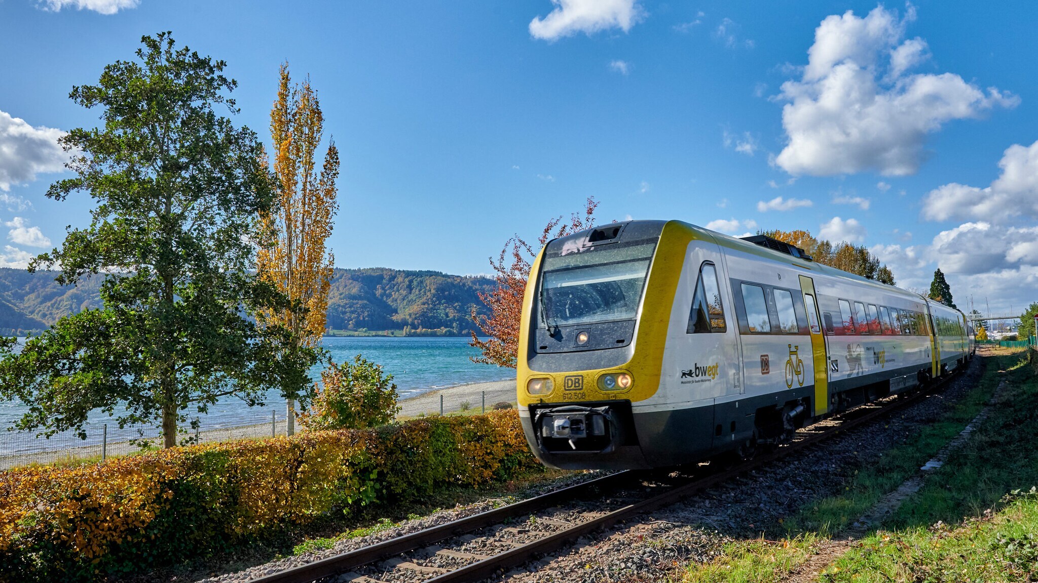 Ein gelb-grüner Zug der Bodensee-Oberschwaben-Bahn fährt entlang der Gleise am Bodensee. Im Hintergrund sind Bäume und Hügel sichtbar.