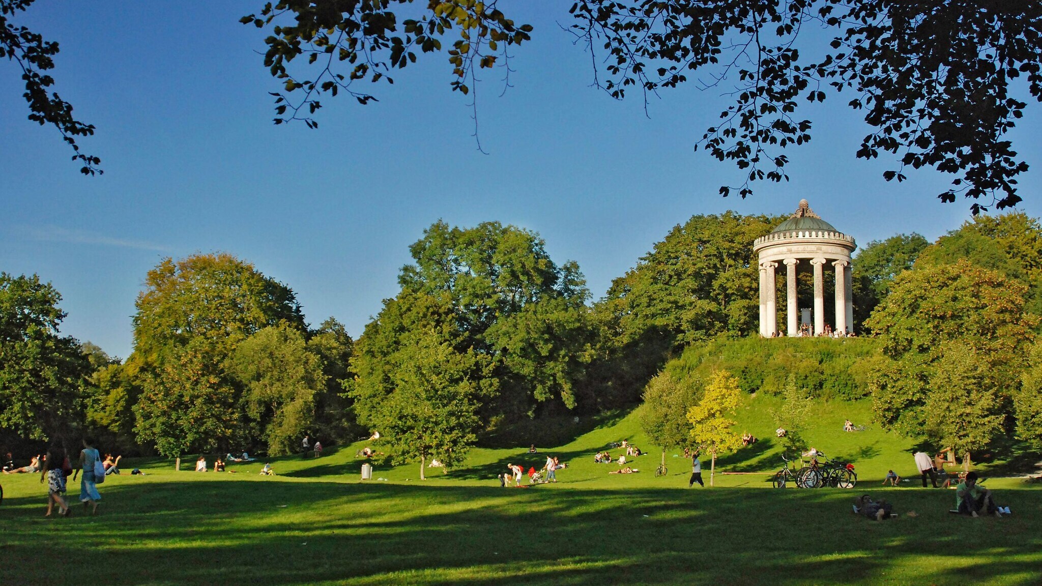 Monopteros-Tempel auf einem Hügel im Englischen Garten in München, umgeben von grünen Bäumen und Wiesen mit Menschen, die sich entspannen