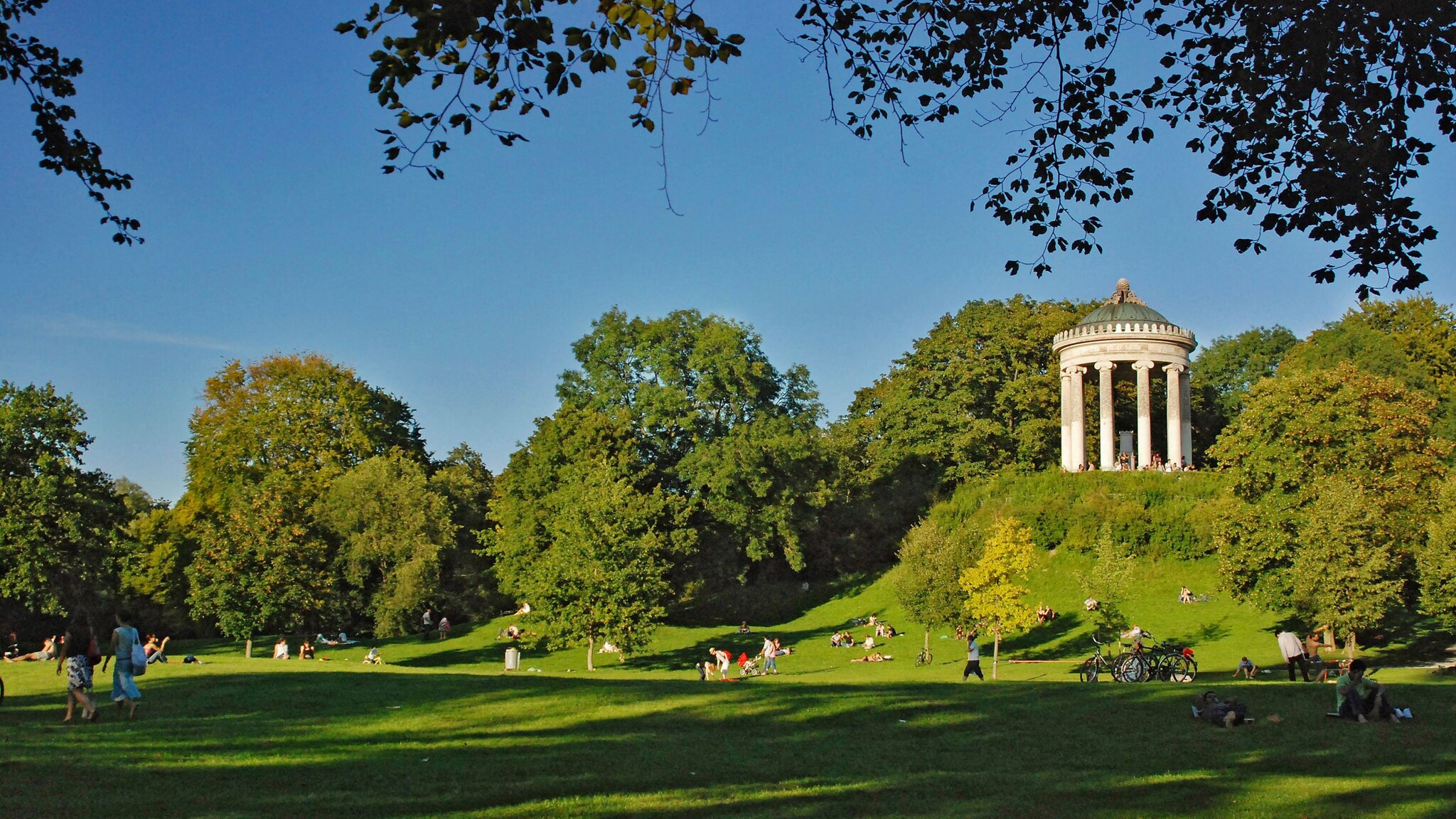 Monopteros-Tempel auf einem Hügel im Englischen Garten in München, umgeben von grünen Bäumen und Wiesen mit Menschen, die sich entspannen Monopteros-Tempel auf einem Hügel im Englischen Garten in München, umgeben von grünen Bäumen und Wiesen mit Menschen, die sich entspannen