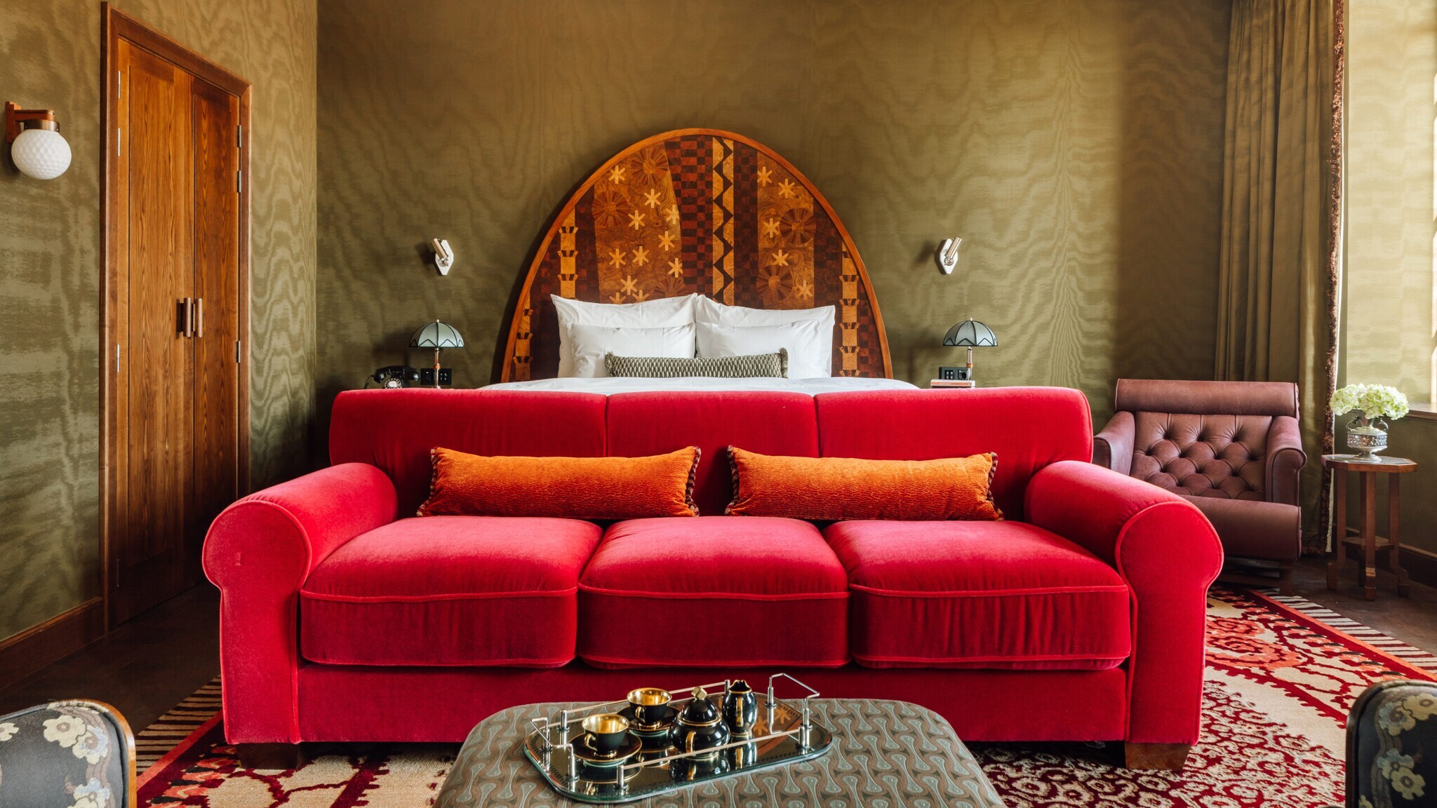 Rotes Sofa mit orangefarbenem Kissen vor einem Bett mit rundem, gemustertem Kopfteil in einem Raum mit grünen Wänden und braunen Holztüren. Rotes Sofa mit orangefarbenem Kissen vor einem Bett mit rundem, gemustertem Kopfteil in einem Raum mit grünen Wänden und braunen Holztüren.