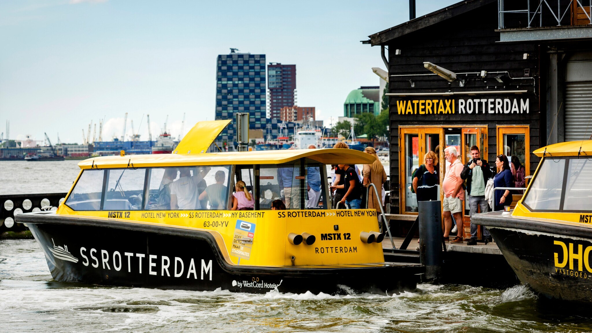 Gelber Wassertaxi-Boot mit der Aufschrift Gelber Wassertaxi-Boot mit der Aufschrift 'SS ROTTERDAM' auf dem Wasser vor einer Anlegestelle mit Schild 'WATERTAXI ROTTERDAM' und mehreren wartenden Personen.