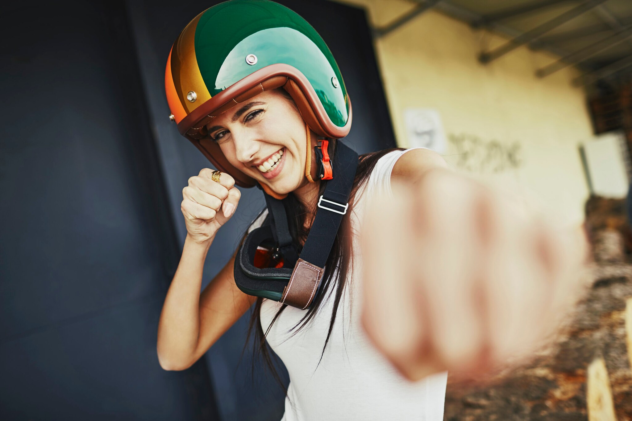 Eine junge, lachende Frau mit einem bunten Motorradhelm auf dem Kopf boxt spielerisch in Richtung Kamera