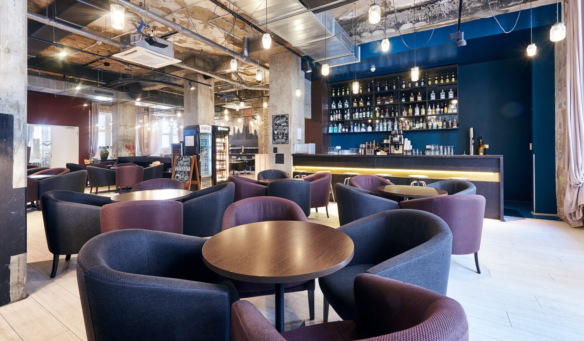 Eine Hotellobby mit runden Tischen und Sesseln, im Hintergrund eine Bar, an der Decke hängen Glühbirnen an Kabeln Eine Hotellobby mit Sitzgelegenheiten vor einer Bar unter einer offenen Raumdecke im Industriestil.