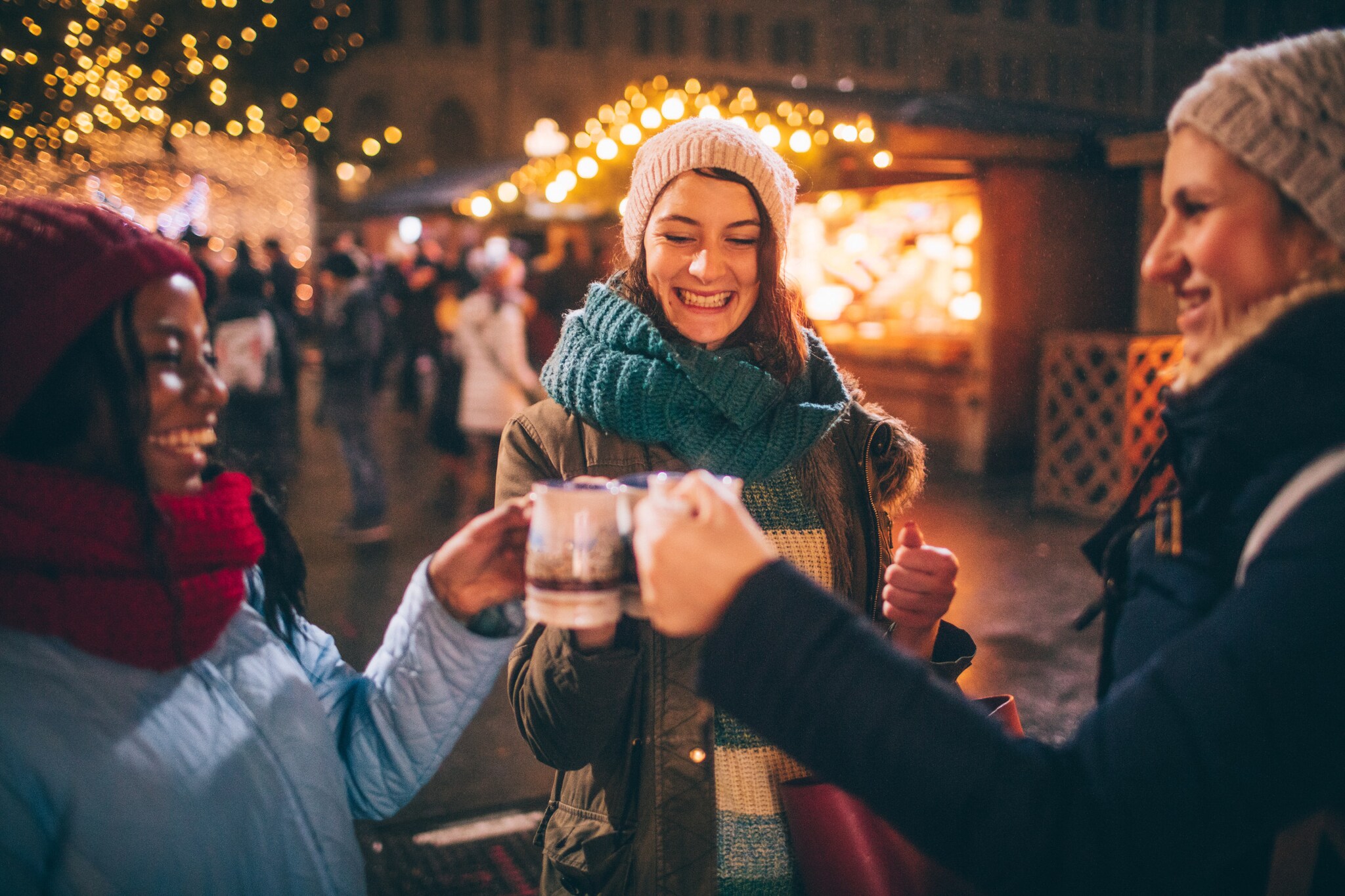 Drei Frauen in Winterkleidung stoßen mit Tassen auf einem Weihnachtsmarkt am Abend an.