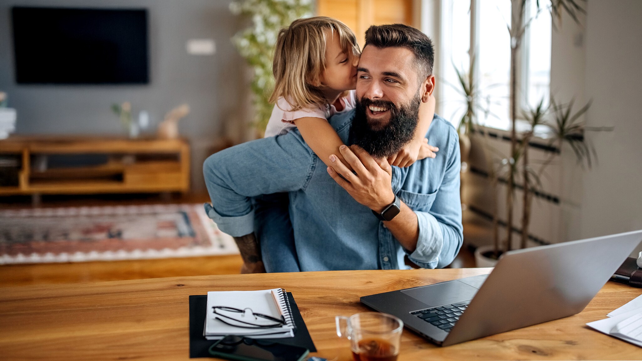 Ein Kind umarmt einen lachenden Mann, der an einem Tisch mit Laptop in einem Wohnzimmer sitzt.