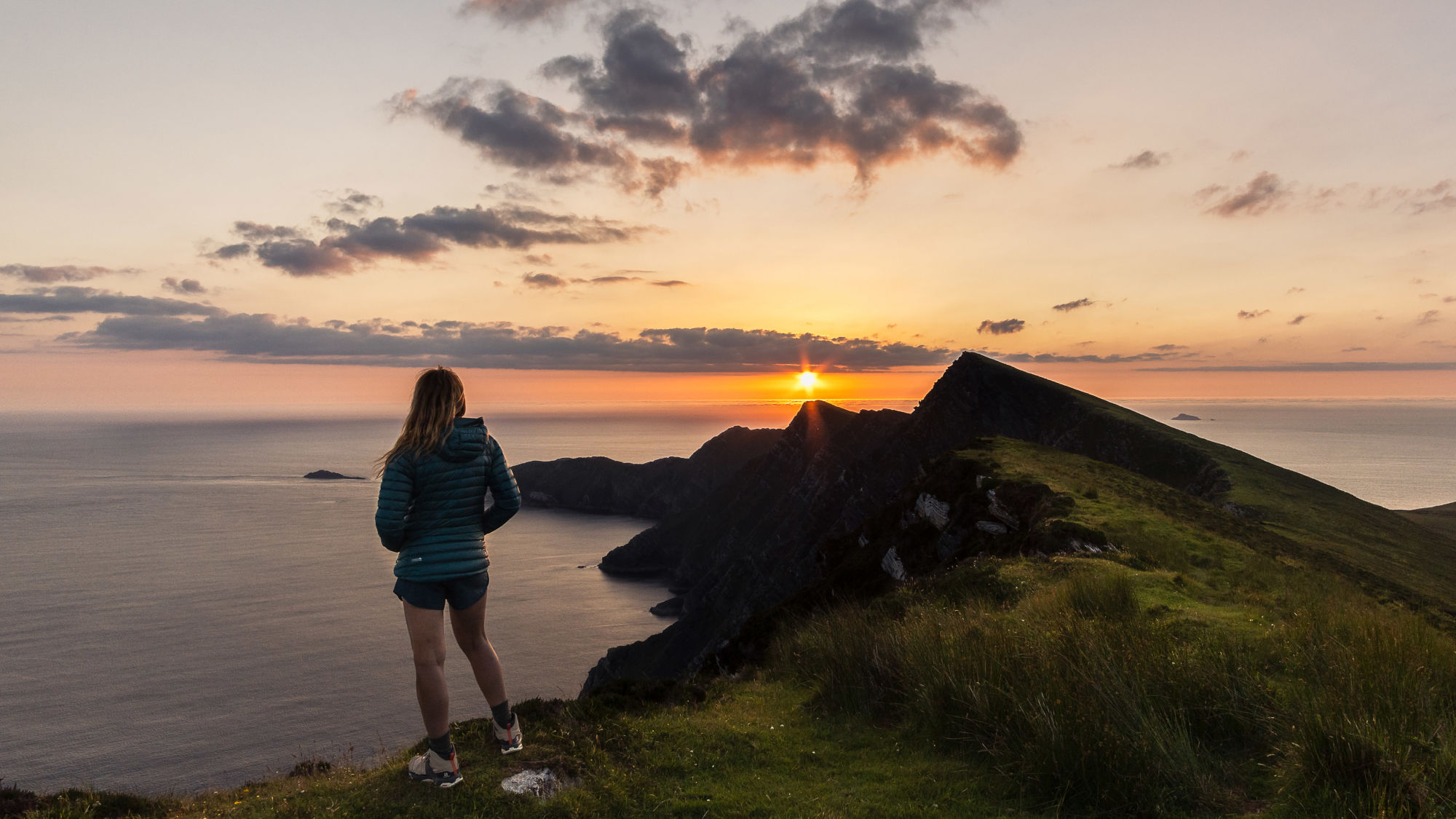 Eine Frau mit langen Haaren steht auf einem Hügel und schaut auf einen Sonnenuntergang über dem Meer. Sie trägt eine grüne Jacke und Shorts. Im Hintergrund sind dunkle Felsen und der Horizont sichtbar. Eine Frau mit langen Haaren steht auf einem Hügel und schaut auf einen Sonnenuntergang über dem Meer. Sie trägt eine grüne Jacke und Shorts. Im Hintergrund sind dunkle Felsen und der Horizont sichtbar.